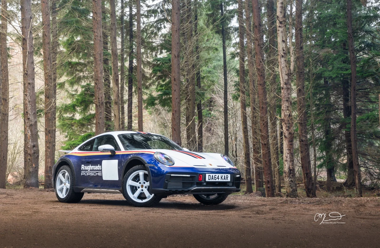 A blue and white Porsche 911 Dakar Roughroads sports car with racing stripes parked on a dirt path in a forested area.