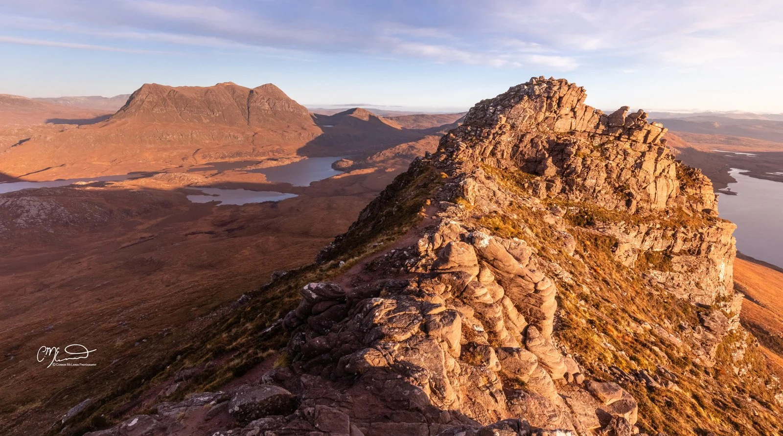 Scotland, north coast 500, NC500, Assynt. Stac Pollaidh. A rocky mountain ridge overlooking a landscape with multiple lakes and distant mountains during sunset.