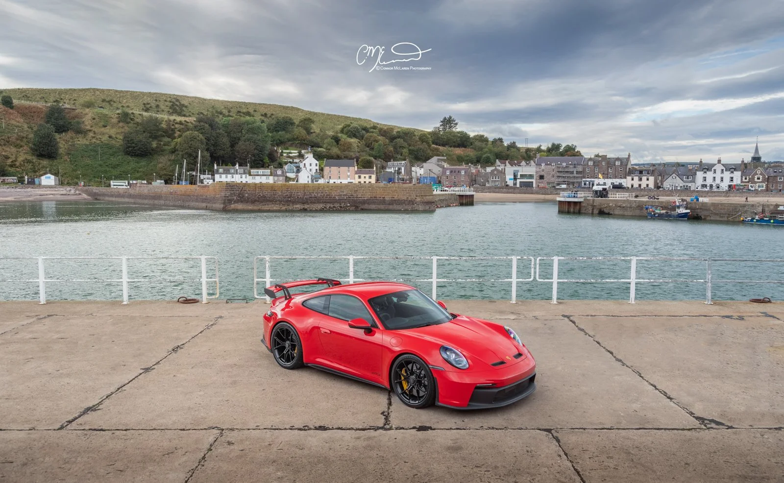 Red Porsche 911 GT3, 992 sports car parked on a concrete pier with a harbor and small town behind it, overlooking water and green hills under a cloudy sky.