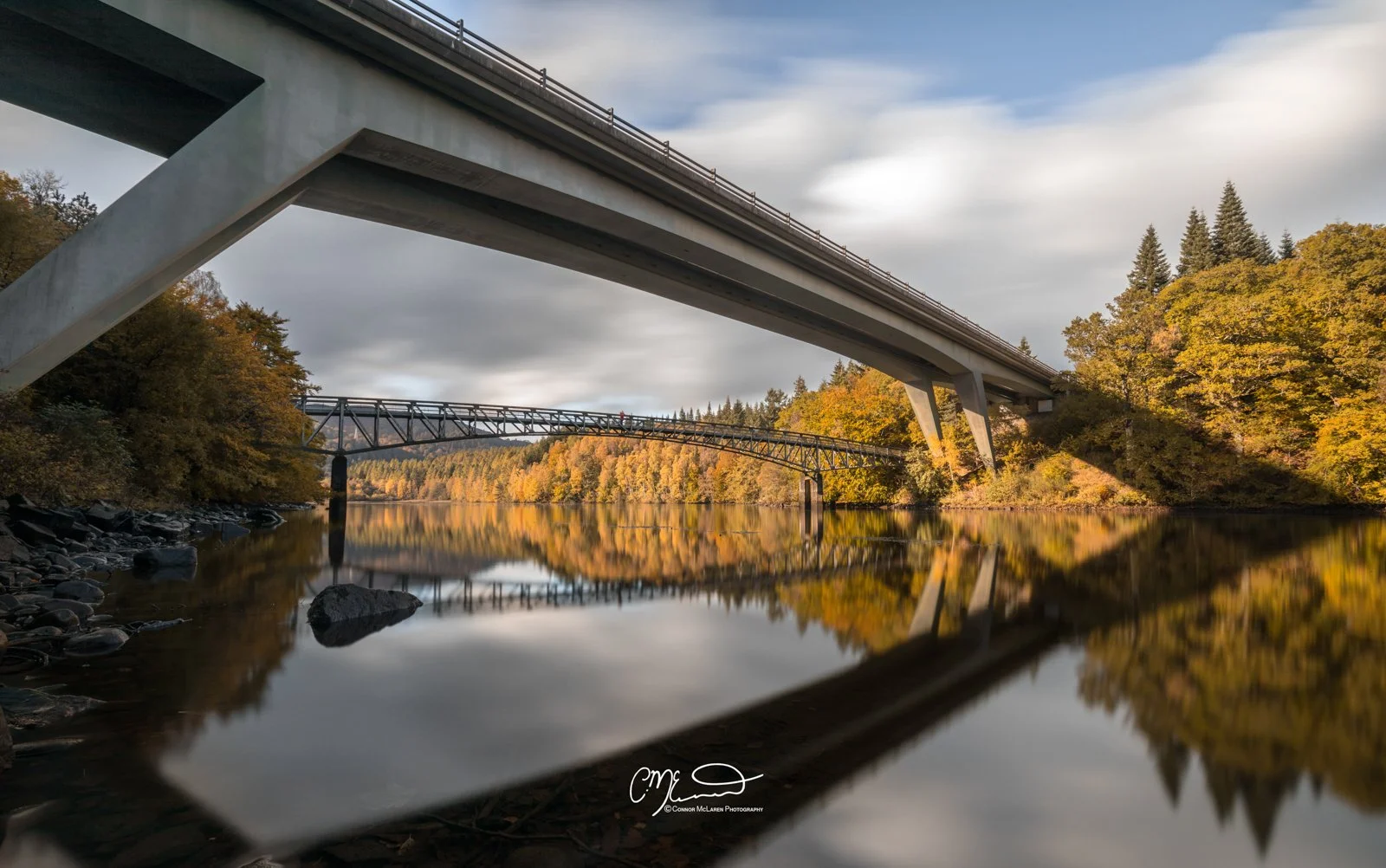 Scottish Central Highlands, Scotland, Perthshire Pitlochry. A river reflecting trees with autumn foliage under a large highway bridge with a second smaller bridge in the background.