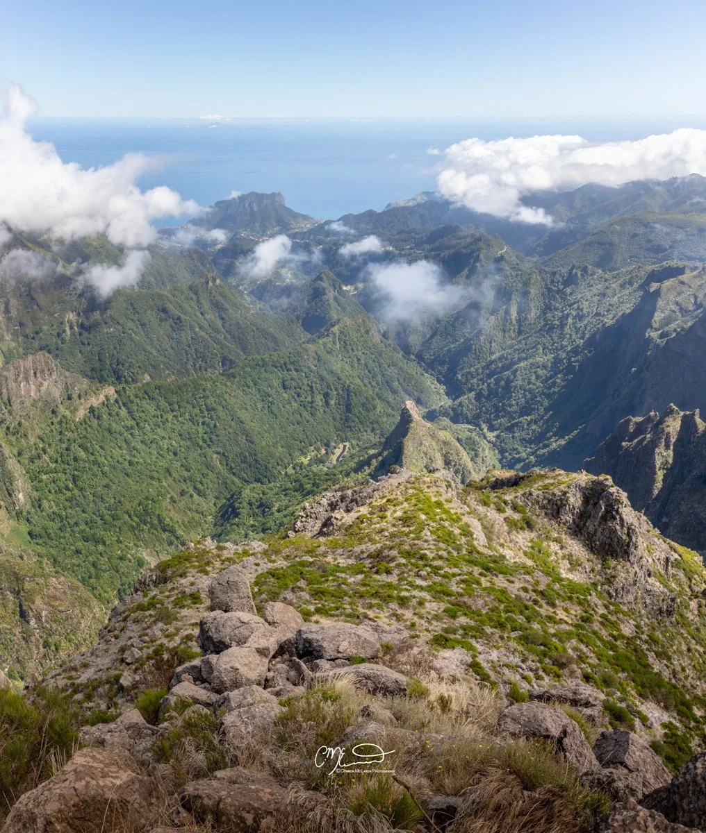 mountain top, Madeira Portugal. Mountain valley landscape with green, rugged mountains and clouds, overlooking the ocean in the distance, rocky foreground with sparse vegetation.