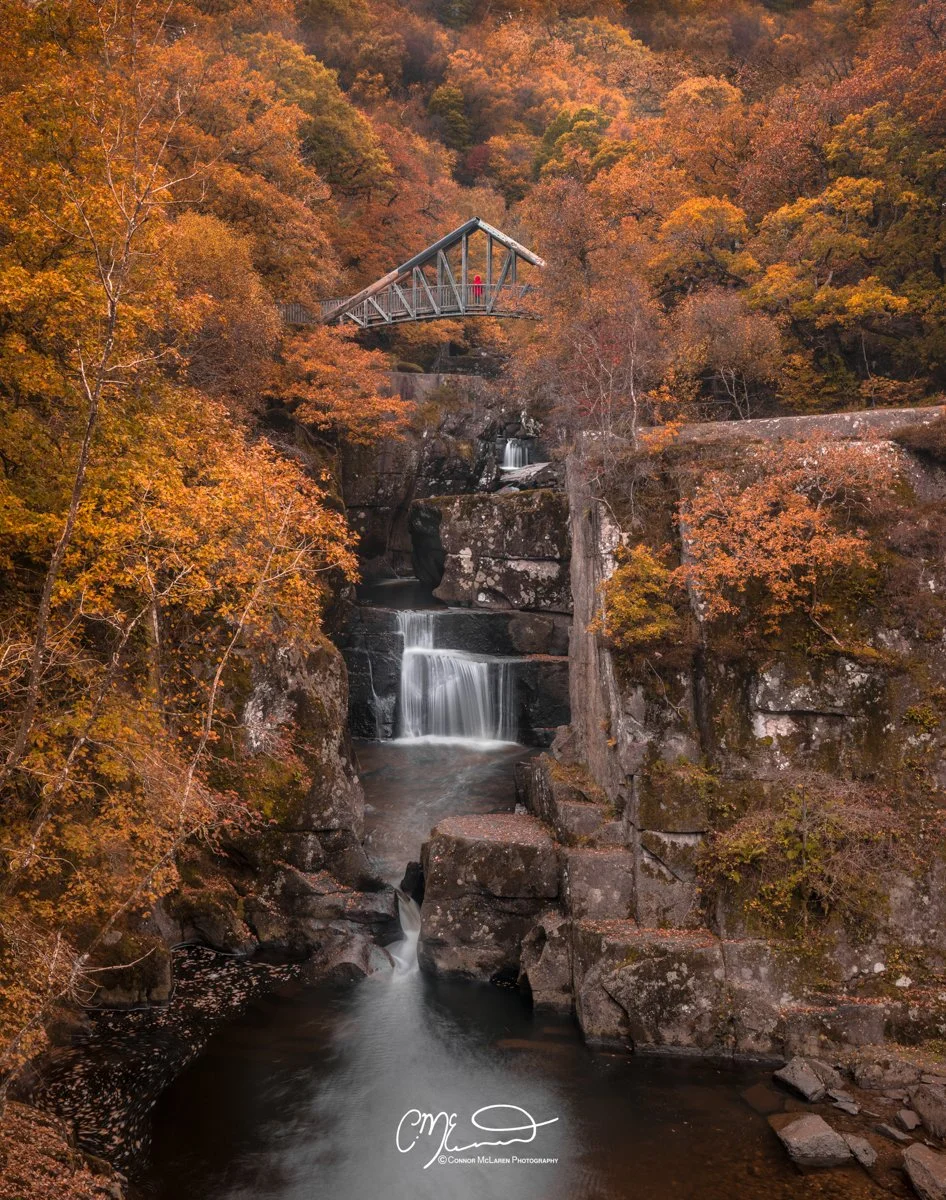 Scottish Lowlands, Scotland, Bracklin Falls. A scenic view of a waterfall cascading down rocks surrounded by trees with autumn-colored leaves. A bridge spans over the gorge with a person standing on it.