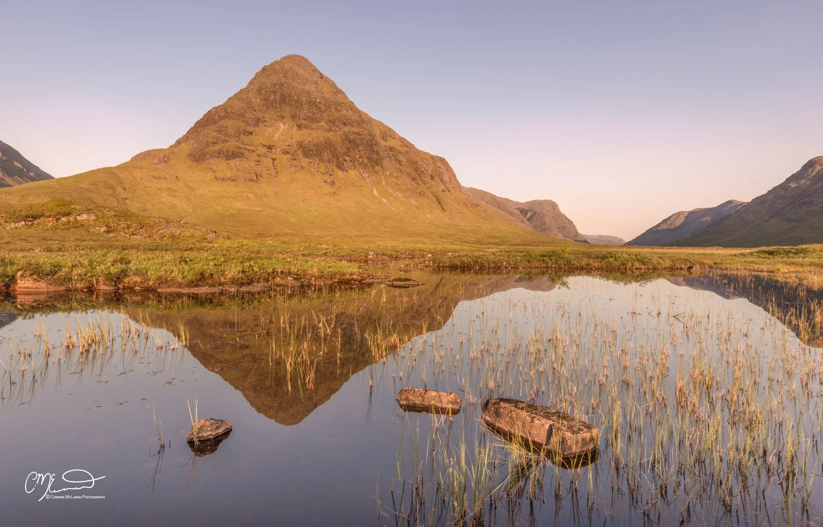 Glen Coe and Glen Etive, Western Highlands, Scotland. Scenic landscape of a mountain with a reflection in a calm body of water, surrounded by grassy terrain and rocks, during sunset or sunrise.