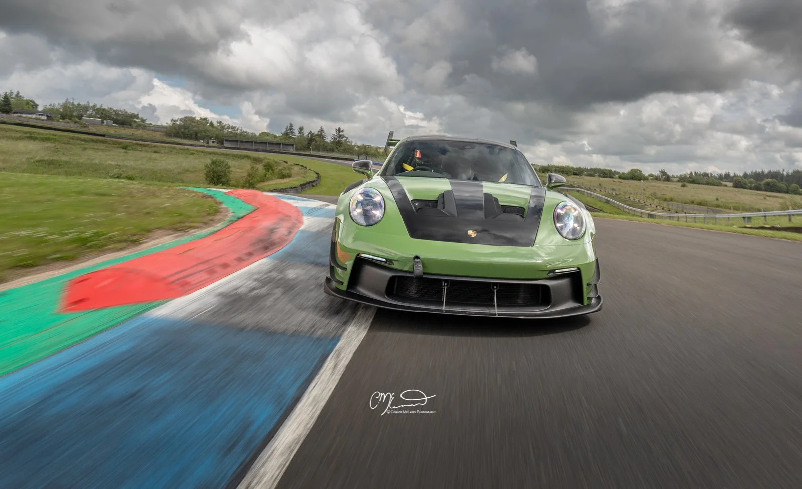 Porsche 911 GT3 RS with Manthey Racing kit in PTS Olive Green, Knockhill racing. Amazing photograph, wallpaper. Green race car on a race track with colorful curbs, under a cloudy sky.