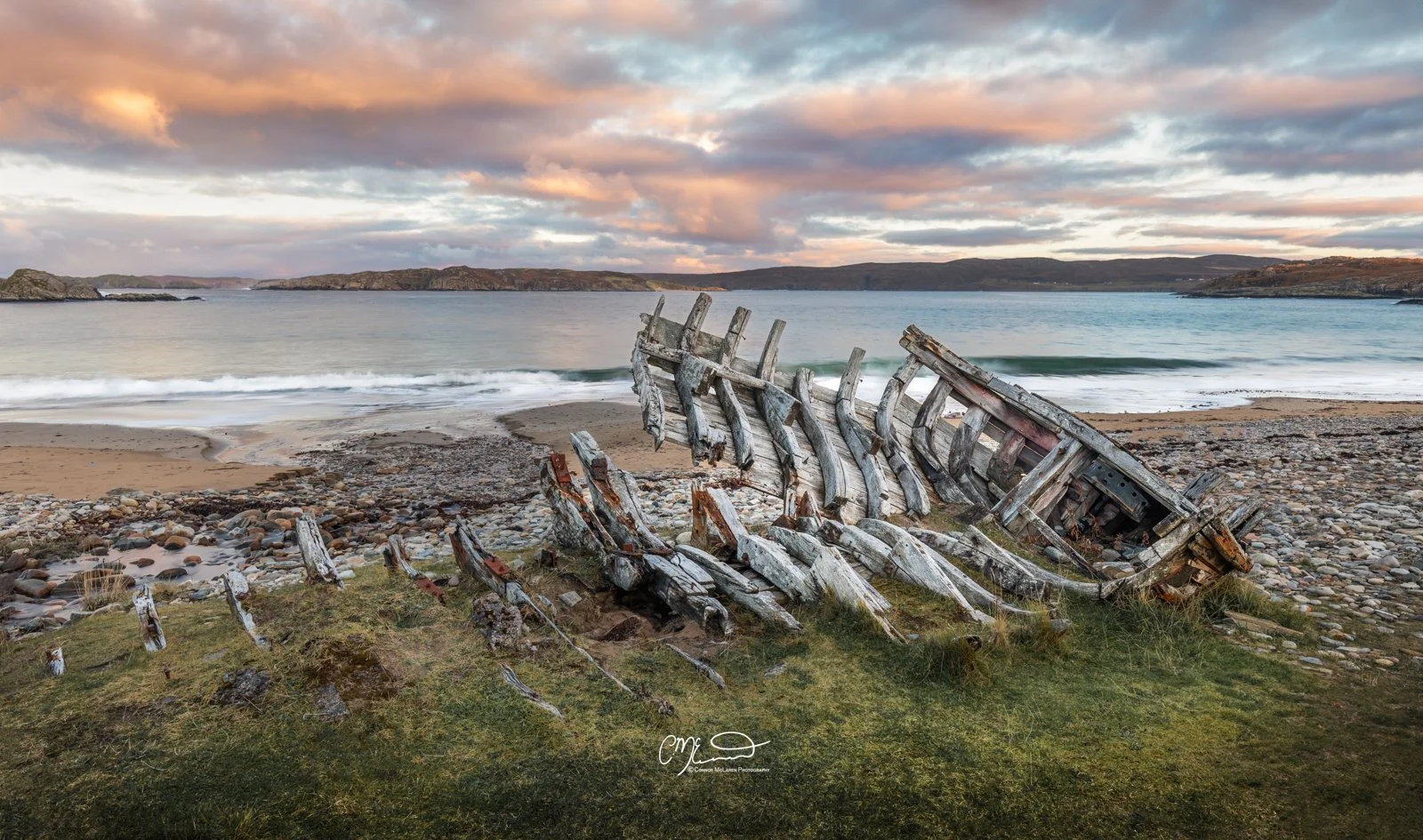 Scotland, north coast 500, NC500. An abandoned, weathered wooden boatwreck on a rocky shoreline with grassy patches, in front of a calm lake and distant hills under a cloudy sky during sunset.