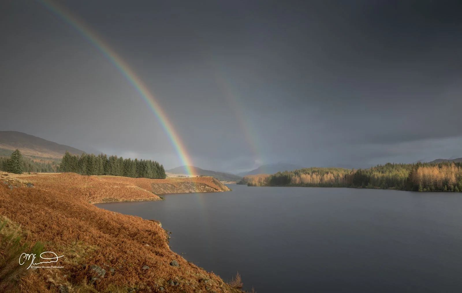 Scottish Central Highlands, Scotland, Perthshire rainbow. A scenic landscape with a river, trees, and hills, under a dark sky with a rainbow.
