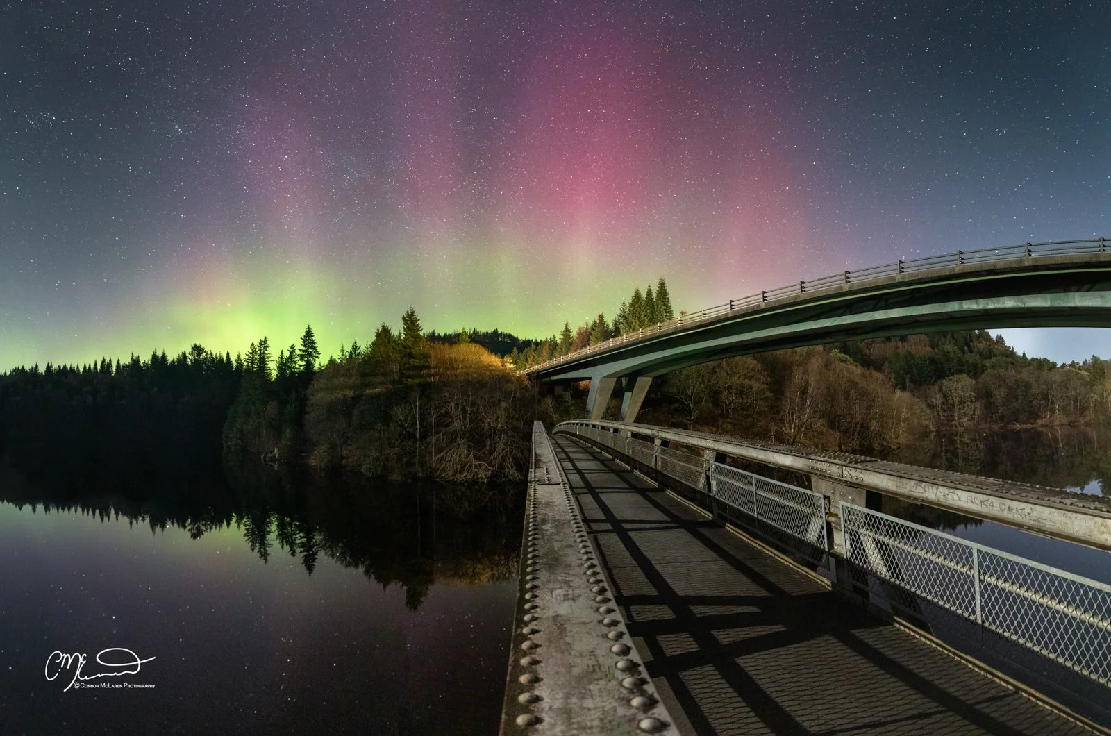 Aurora Northern Lights, Perthshire, Night Sky, Scotland. Night scene showing a bridge over a calm lake reflecting the Northern Lights and starry sky, with trees on hills in the background.