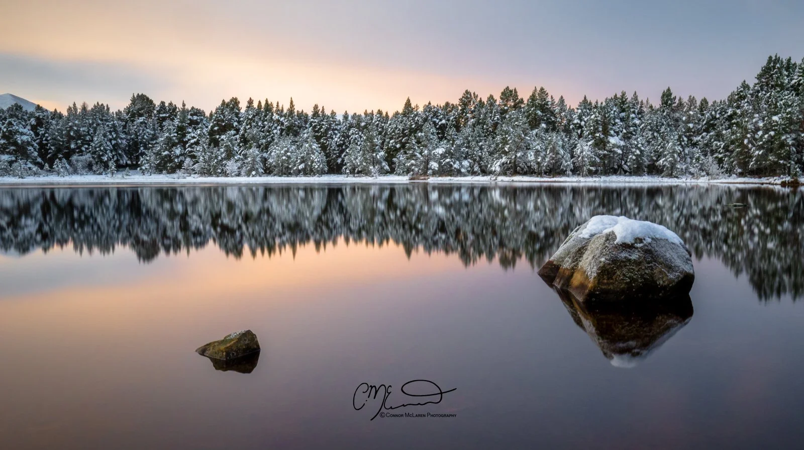 Scottish Central Highlands, Scotland, Cairngorms, Loch Morlich. . A calm lake reflecting snow-covered trees and a partly cloudy sky during sunset, with rocks partially submerged in the water.