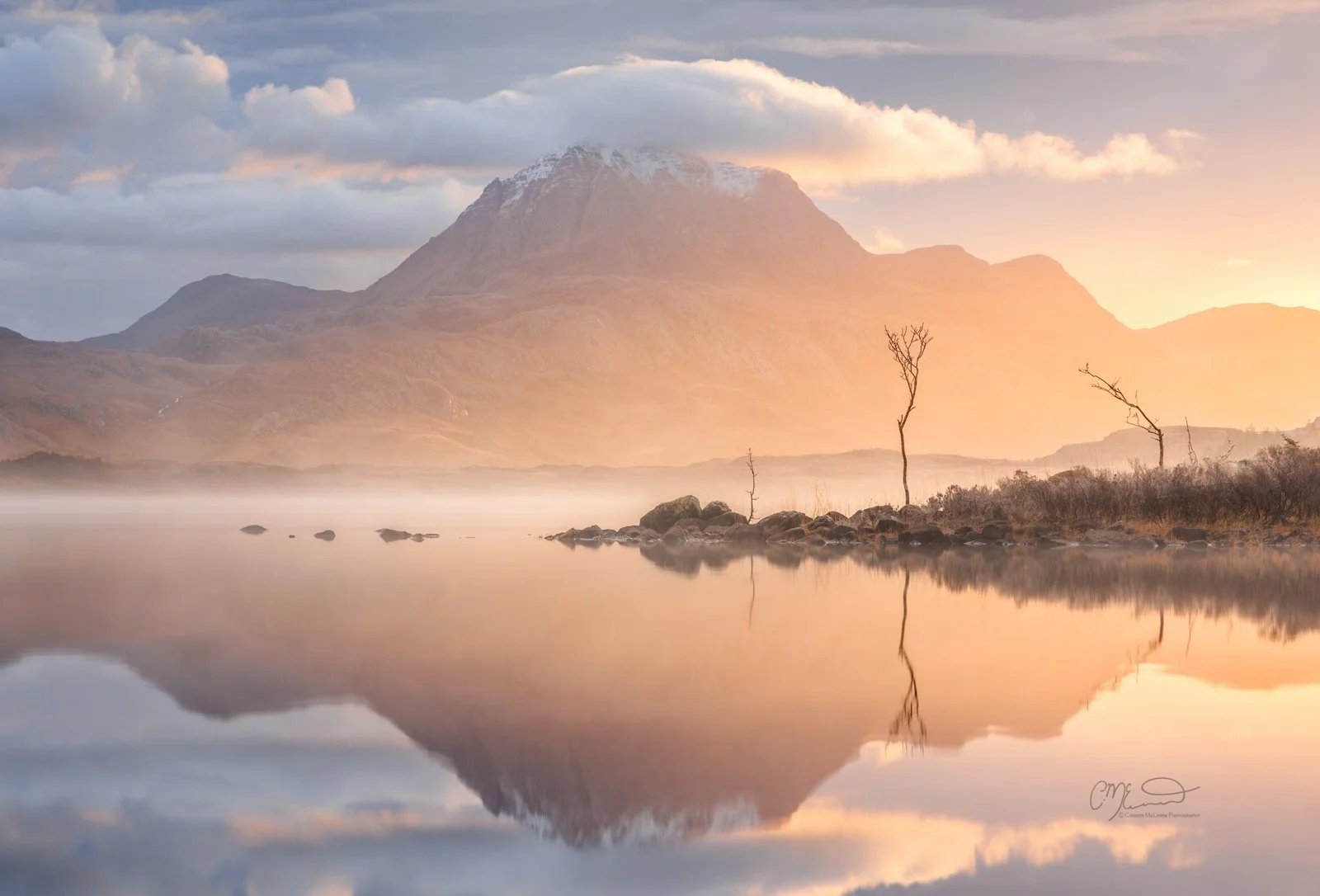 Scotland, north coast 500, NC500, Loch Maree. A serene mountain landscape featuring a tall, snow-capped mountain reflecting in a calm lake, with a few small, leafless trees and rocks along the shoreline.