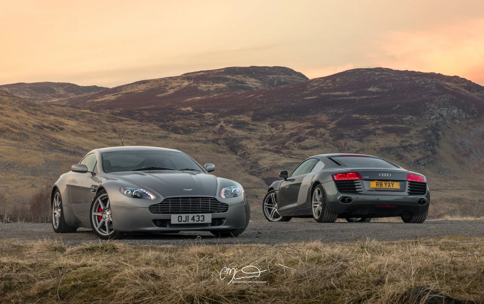Two luxury sports cars, an Aston Martin and an Audi R8, parked on a rural road with rolling hills in the background during sunset.