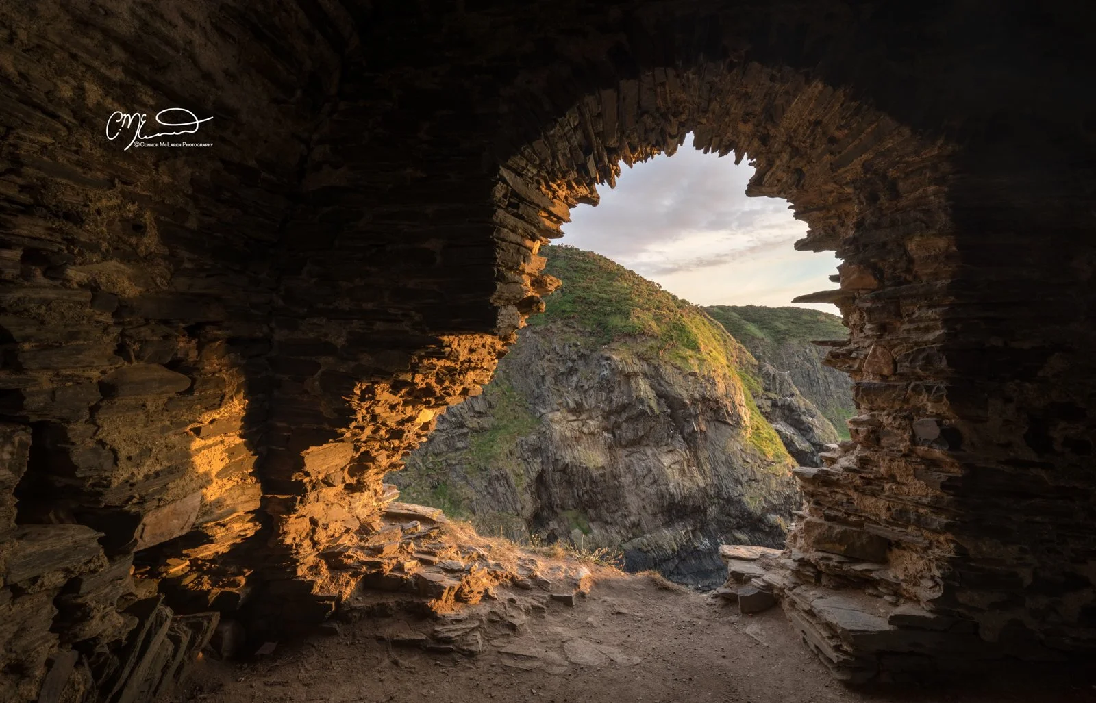 Findlater Castle, Morrayshire, North East Scotland. View through a stone window in a cliffside, overlooking green mountains and a cloudy sky at sunset.