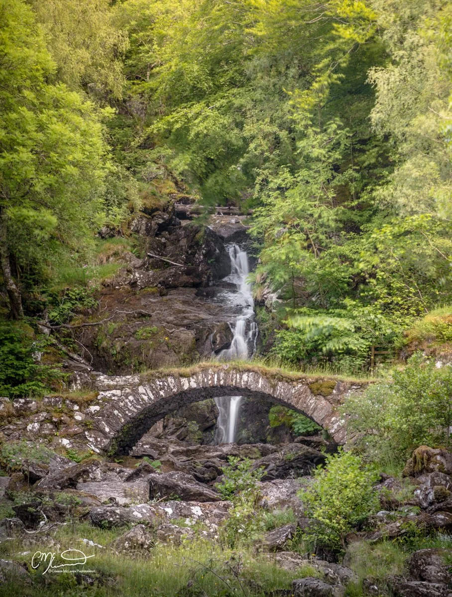 Scottish Central Highlands, Scotland, Perthshire glen lyon. A stone arch bridge crosses a rocky stream with a small waterfall, surrounded by lush green trees.