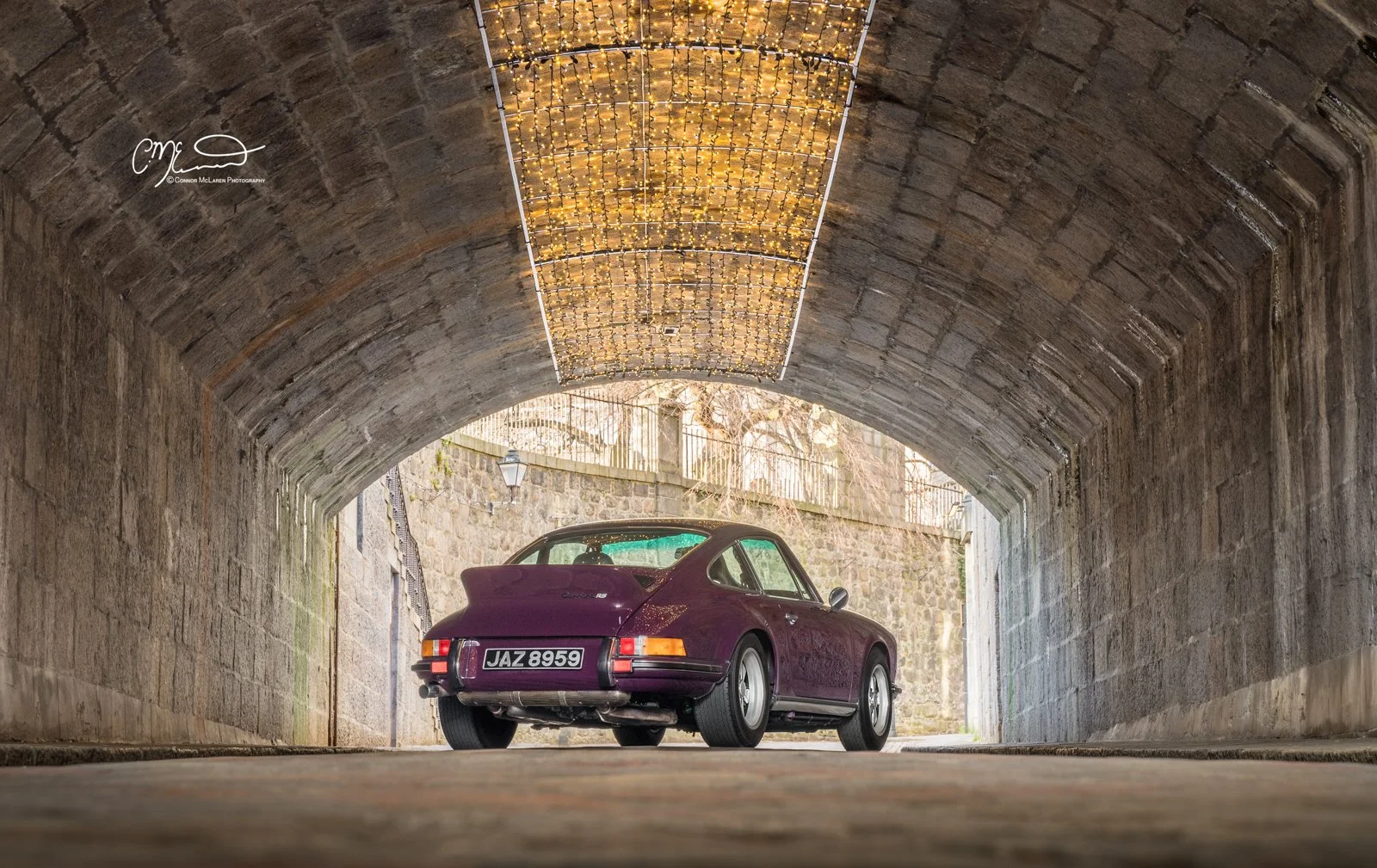 Porsche 911, 2.7RS. A vintage purple Porsche car parked under a stone tunnel with warm overhead string lights.