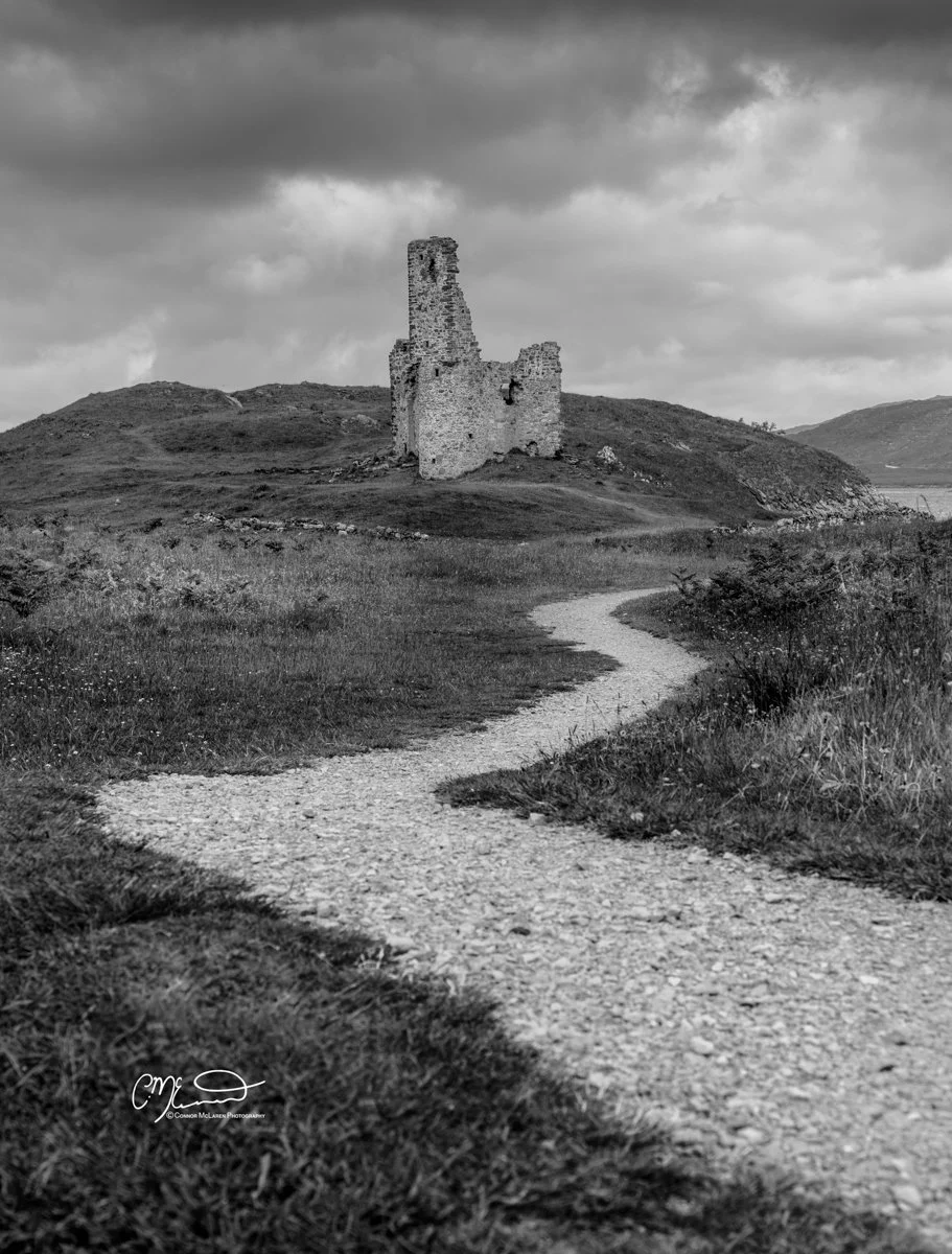 Scotland, Loch Assynt, Ardvrek Castle. Black and white photo of a winding dirt path leading to an old, ruined stone castle on a hill under cloudy skies.