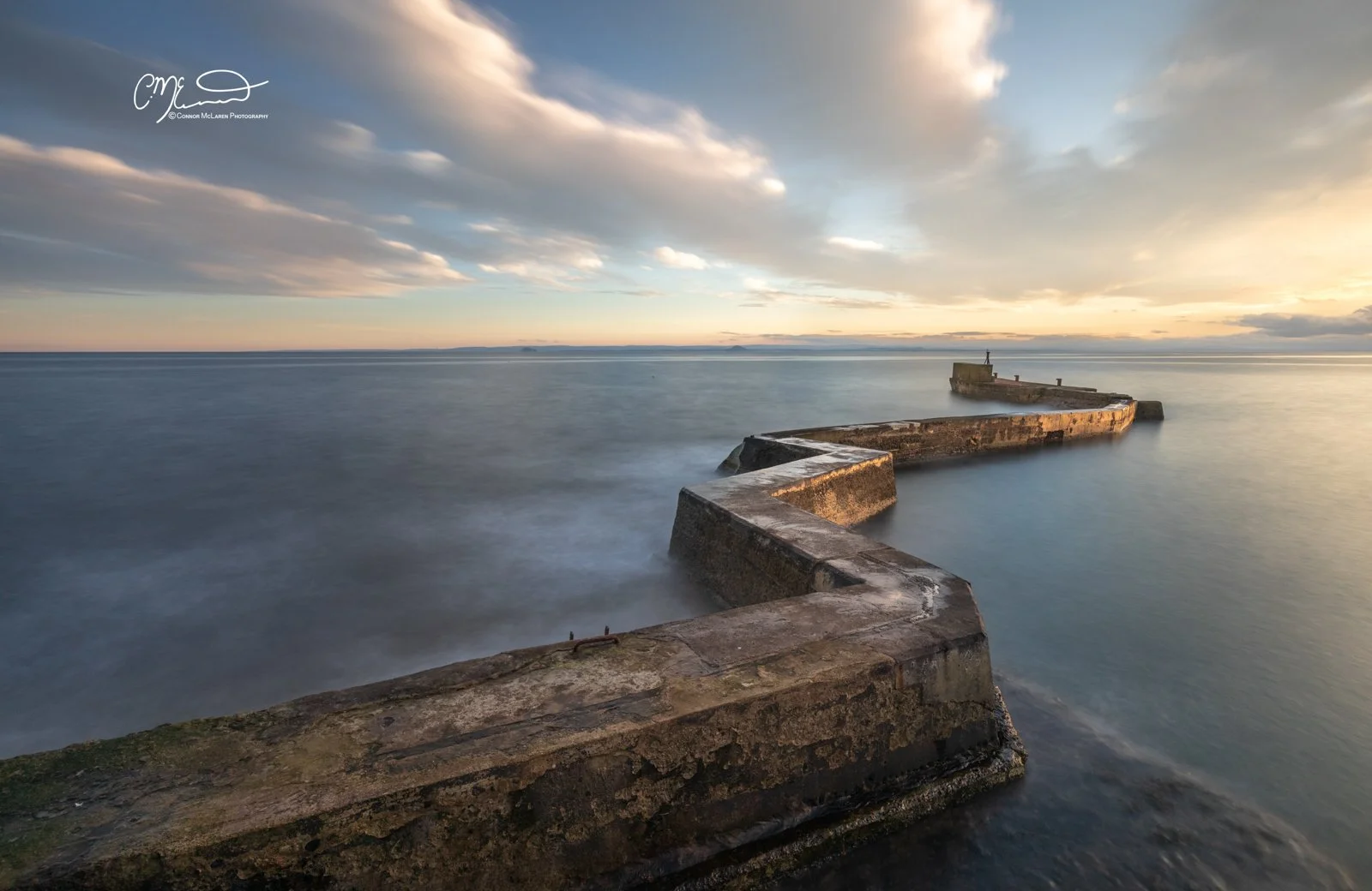 Scottish Lowlands, Scotland, east neuk of fife. A long, zigzag concrete pier extending into calm ocean waters during sunset, with a partly cloudy sky.