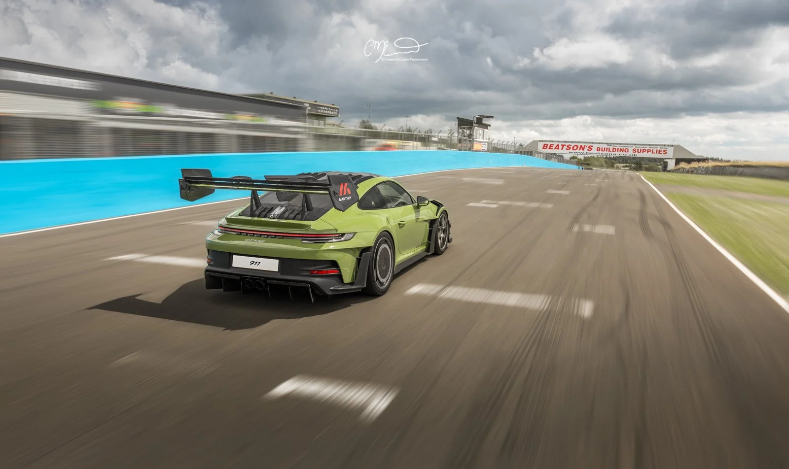 Porsche 911 GT3 RS with Manthey Racing kit in PTS Olive Green, Knockhill racing. Amazing photograph, wallpaper. Green Porsche 911 racing car on a track with cloudy sky, blue barriers, and a sign for Beatson's Building Supplies in the background.