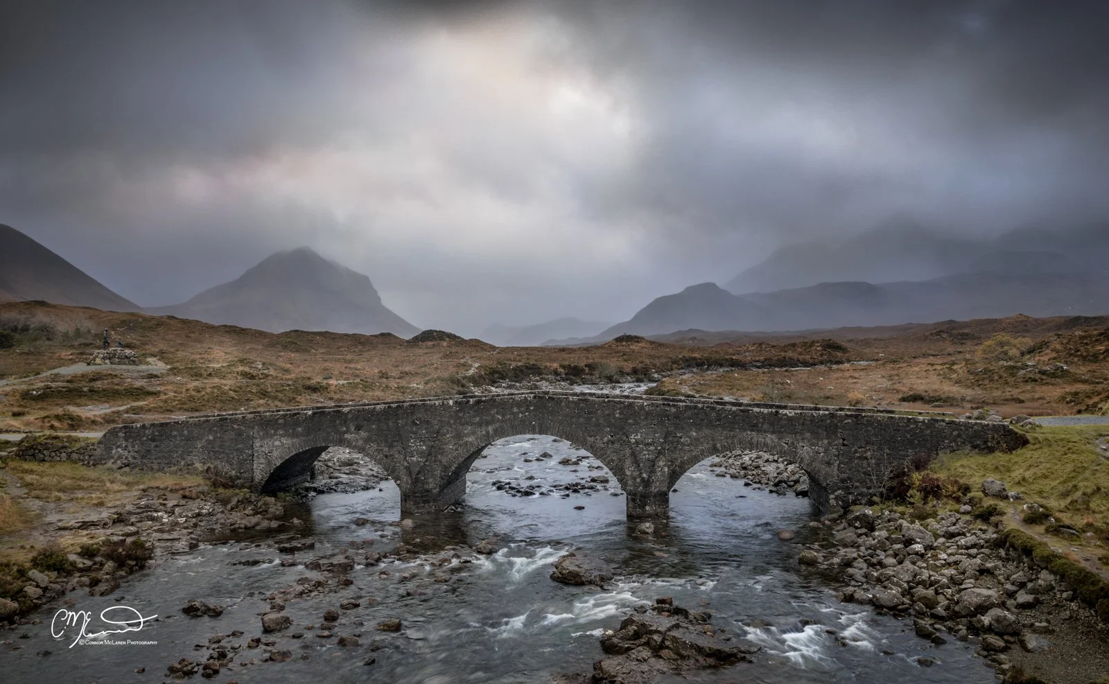 Scotland, Isle of Skye, sligachan. Stone bridge over a flowing river in a mountainous landscape with dark, cloudy sky