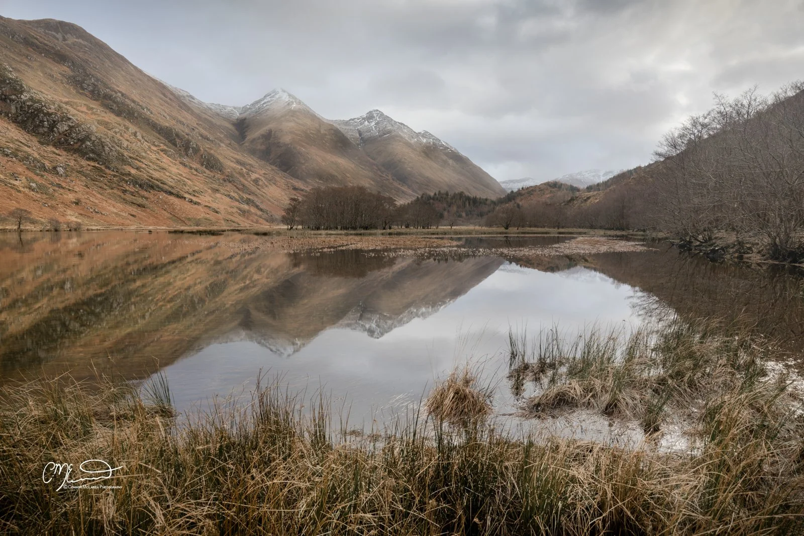 Glen Shiel, Western Highlands, Scotland. Scenic view of a calm lake reflecting the surrounding brown hills, bare trees, and snow-capped mountains under a cloudy sky.