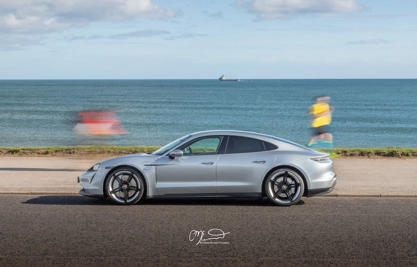 A silver Porsche Taycan car parked beside a seaside road with blurred people walking in the background, and the ocean with a ship in the distance under a partly cloudy sky.