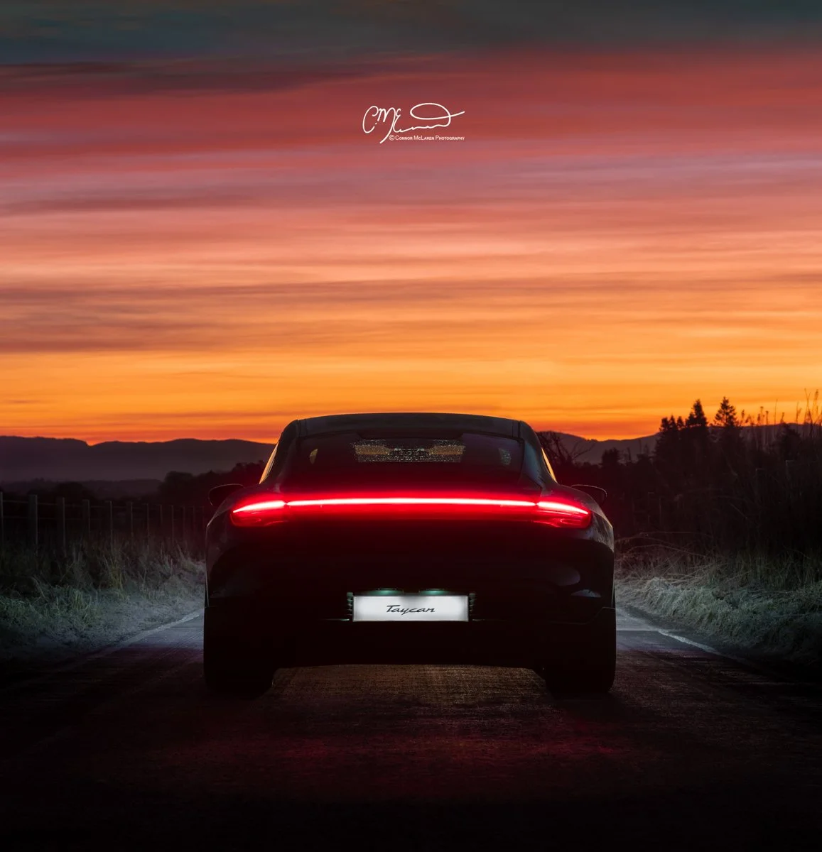 Rear view of a black Porsche Taycan with illuminated red taillights on a rural road at sunset, with a colorful orange, pink, and purple sky and distant mountains in the background.