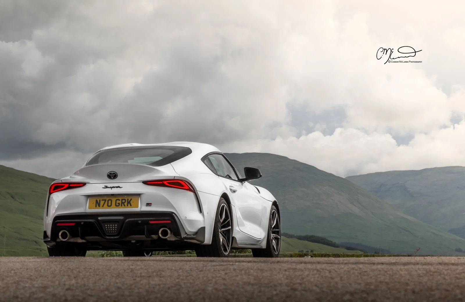 A white Toyota Supra sports car parked on a rural road with green hills and a cloudy sky in the background.
