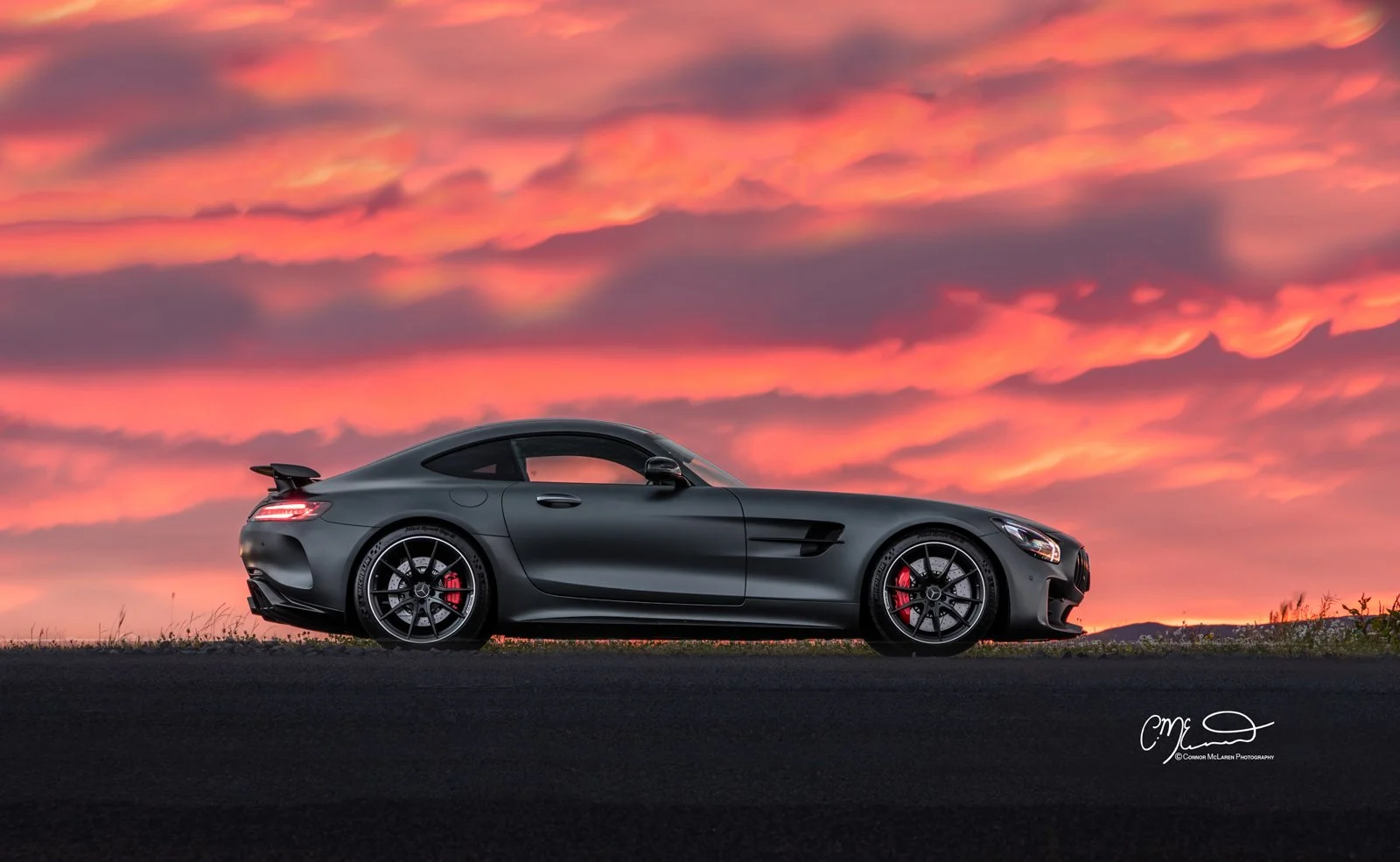 A matte grey Mercedes-benz AMG GTR sports car, likely a Mercedes-Benz AMG GT, parked on a road during sunset with a colorful sky of pink and orange clouds.