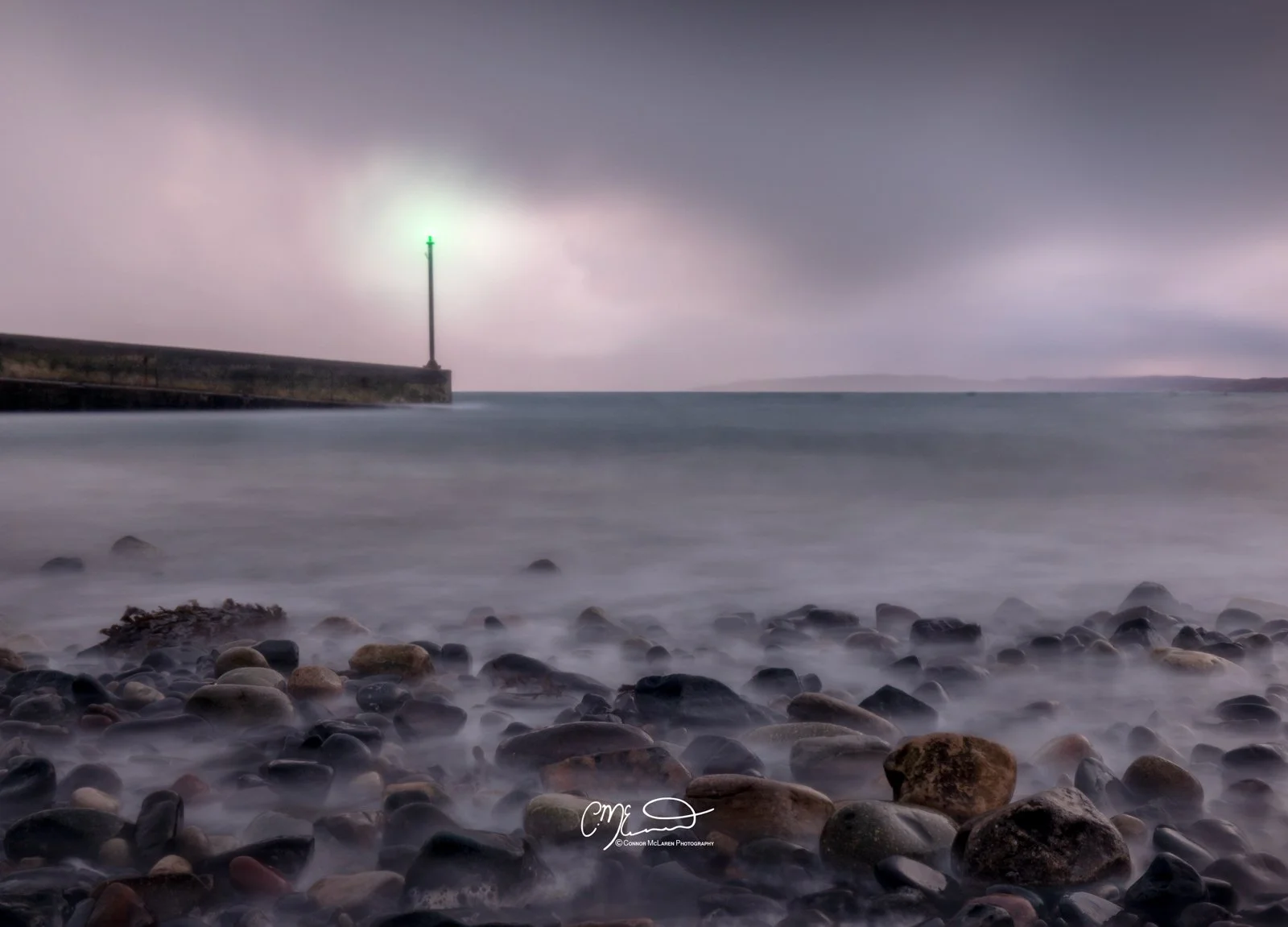 Scotland, Isle of Skye, Elgol. A rocky beach with a lighthouse in the distance, under a cloudy sky.