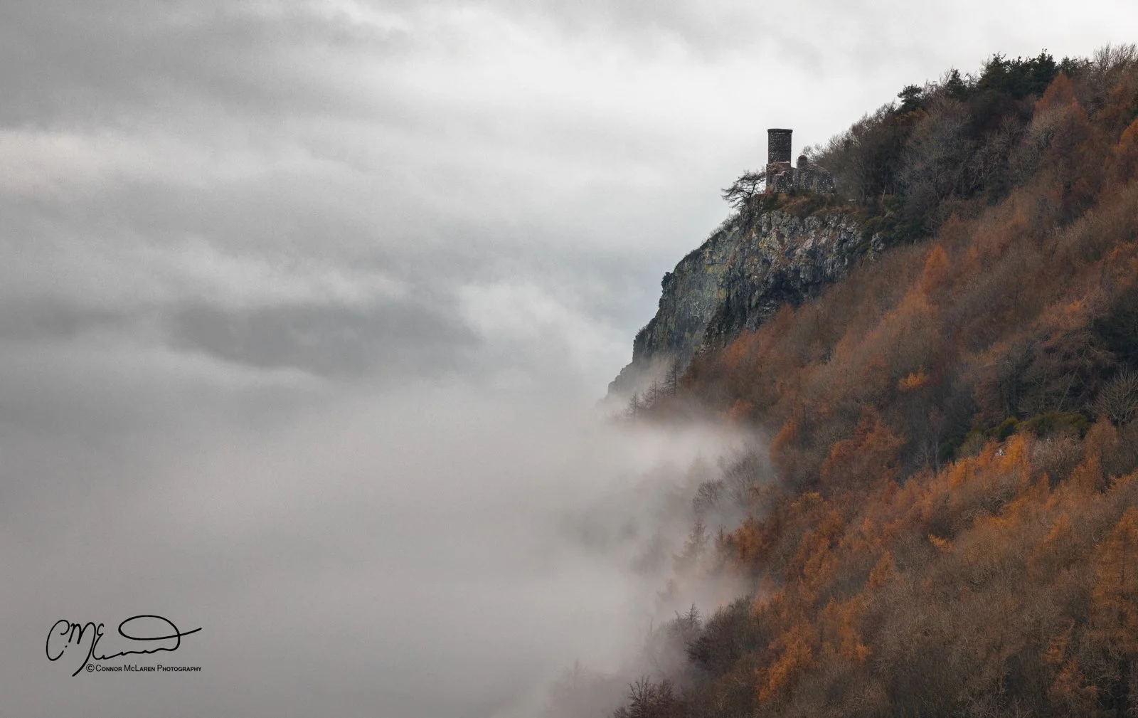 Scottish Central Highlands, Scotland, Perthshire Kinnoull Hill. Cliffside with a small stone building and chimney on top, surrounded by fog and autumn-colored trees along the slope, under a cloudy sky.