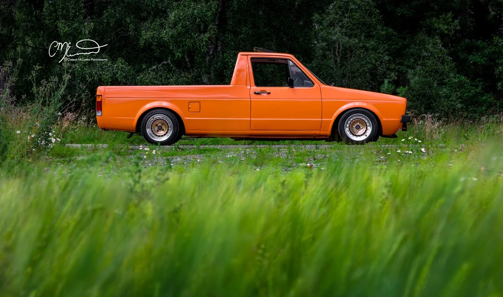 Orange vintage Volkswagen Caddy pickup truck parked on grass with trees in the background.