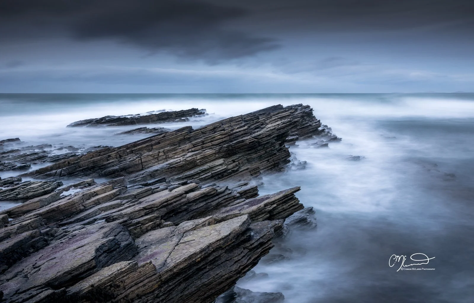 Orkney, Scotland. Long exposure photograph of layered rocks on a stormy ocean shoreline during cloudy weather, with misty waves and a dark sky.