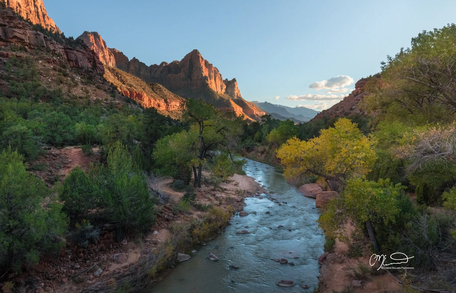 Bucket list locations, USA Zion National Park. A river flowing through a green canyon with trees on both sides and red rock mountains in the background. Clear blue sky with a few clouds.