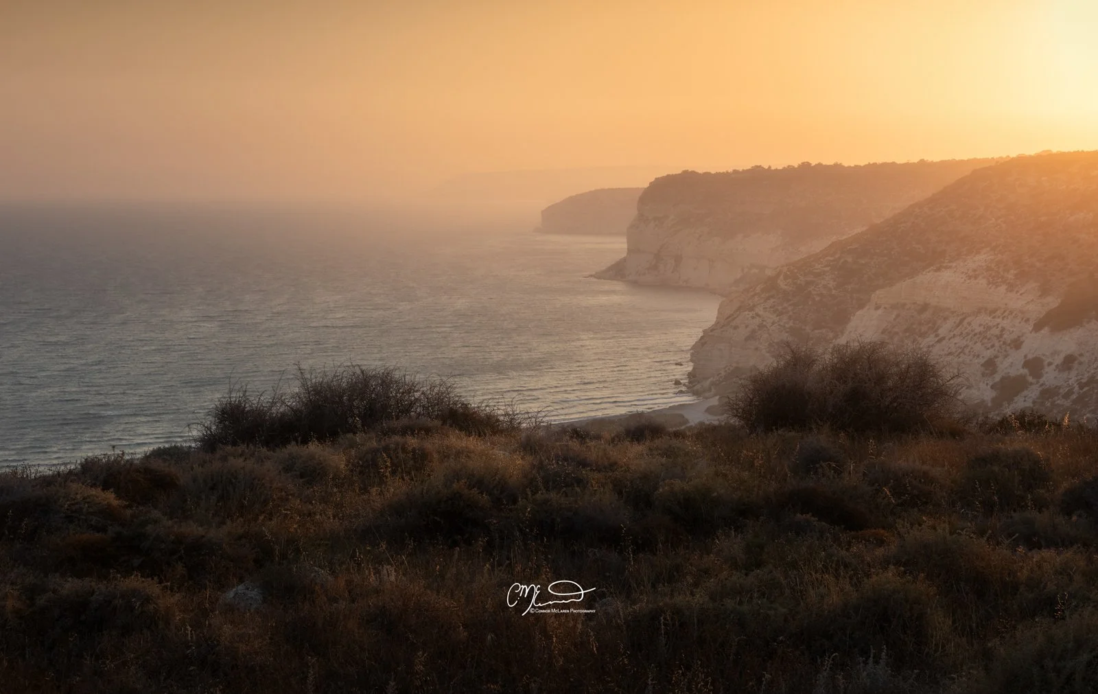 kourion, Cyprus. Sunset over a coastal landscape with cliffs and bushes in the foreground.
