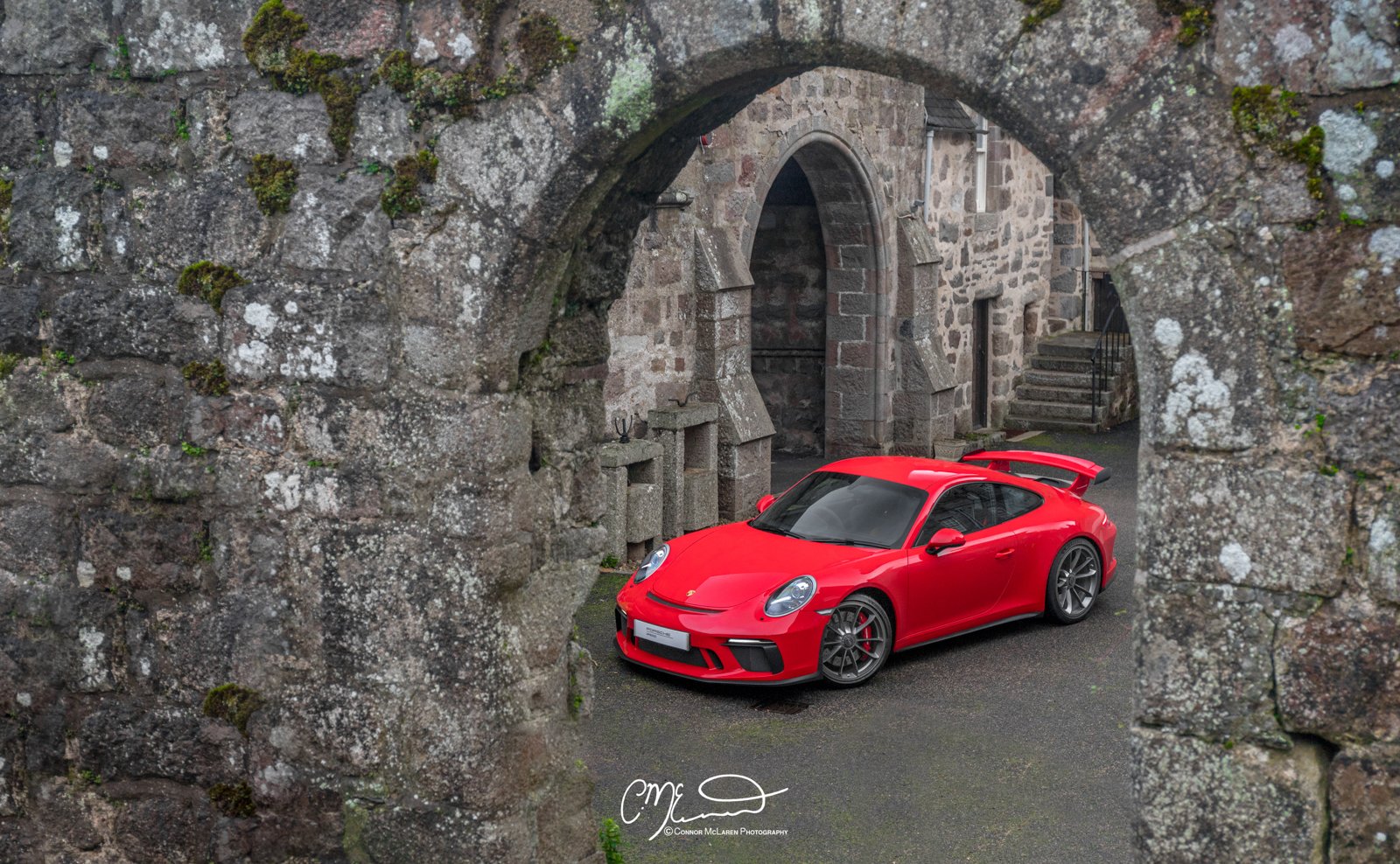 A red Porsche 911 GT3, 991 sports car parked under a stone archway with weathered stone walls and steps in the background.