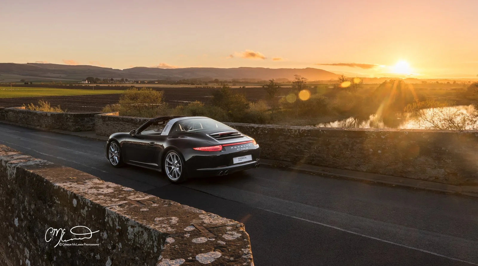 A black Porsche 911 Targa parked on an old stone bridge during sunset with a scenic view of the countryside, fields, trees, and mountains in the background.