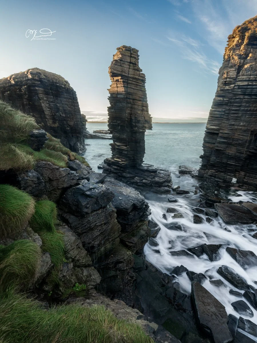 Scotland, north coast 500, NC500, Castle Sinclair Gringoe. Rock formations along a shoreline with water flowing around the rocks, and patches of green grass on the cliffs under a partly cloudy sky.