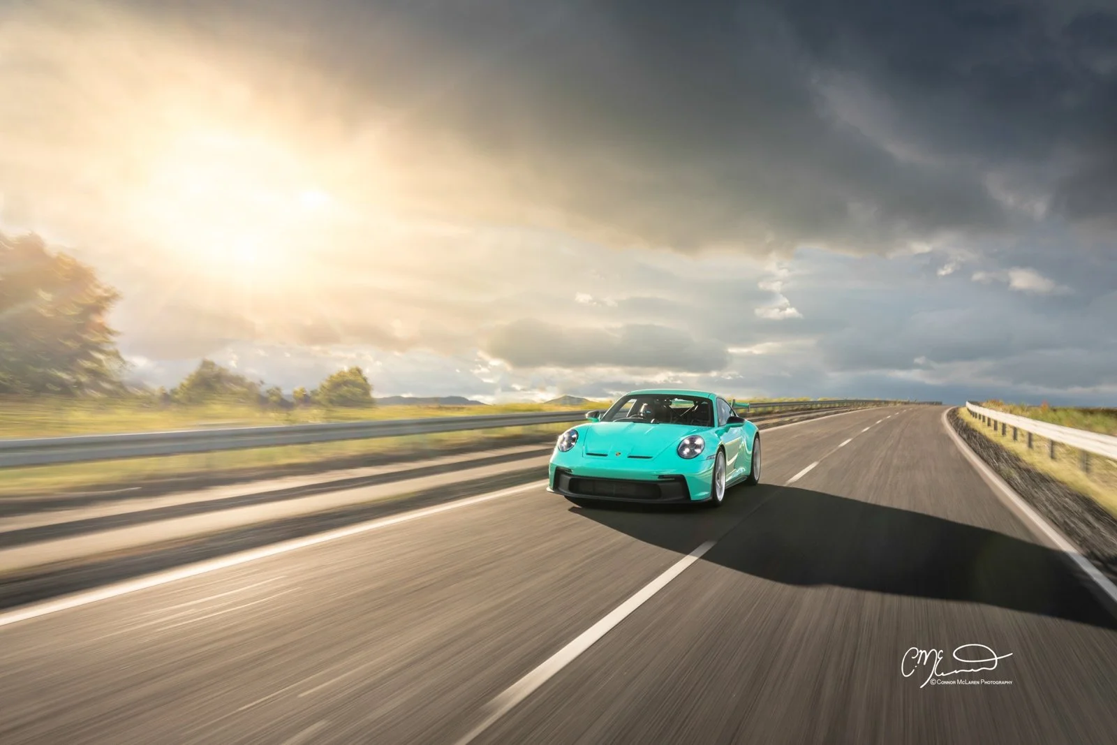 Mint Porsche 911 GT3 992. A light blue sports car driving on a highway with a dramatic cloudy sky and sunlight in the background.