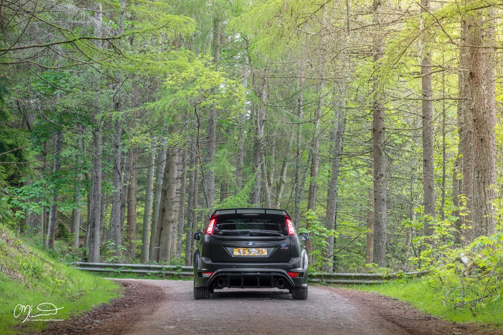 Black Ford Focus RS500 parked on a dirt road in a dense green forest.