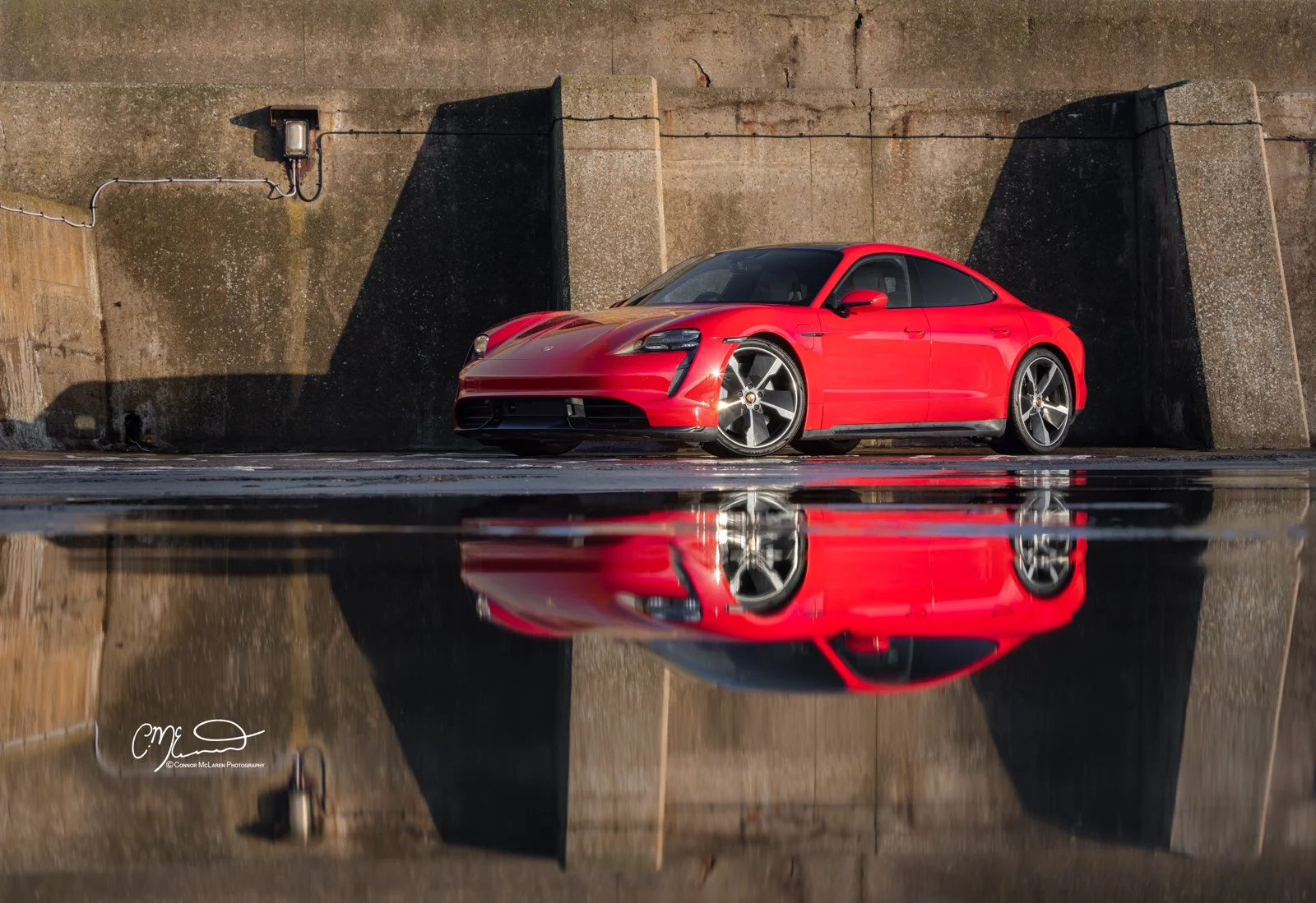 A red Porsche Taycan sports car parked against a concrete wall, with its reflection visible in a puddle of water on the ground.