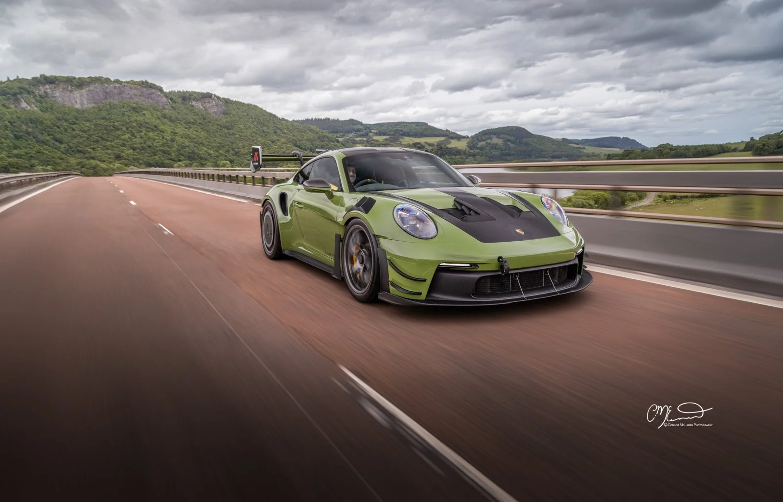 Porsche 911 GT3 RS in Paint to sample Olive green with full Manthey Racing kit. A green Porsche racing car driving on an open highway with mountains and cloudy sky in the background.