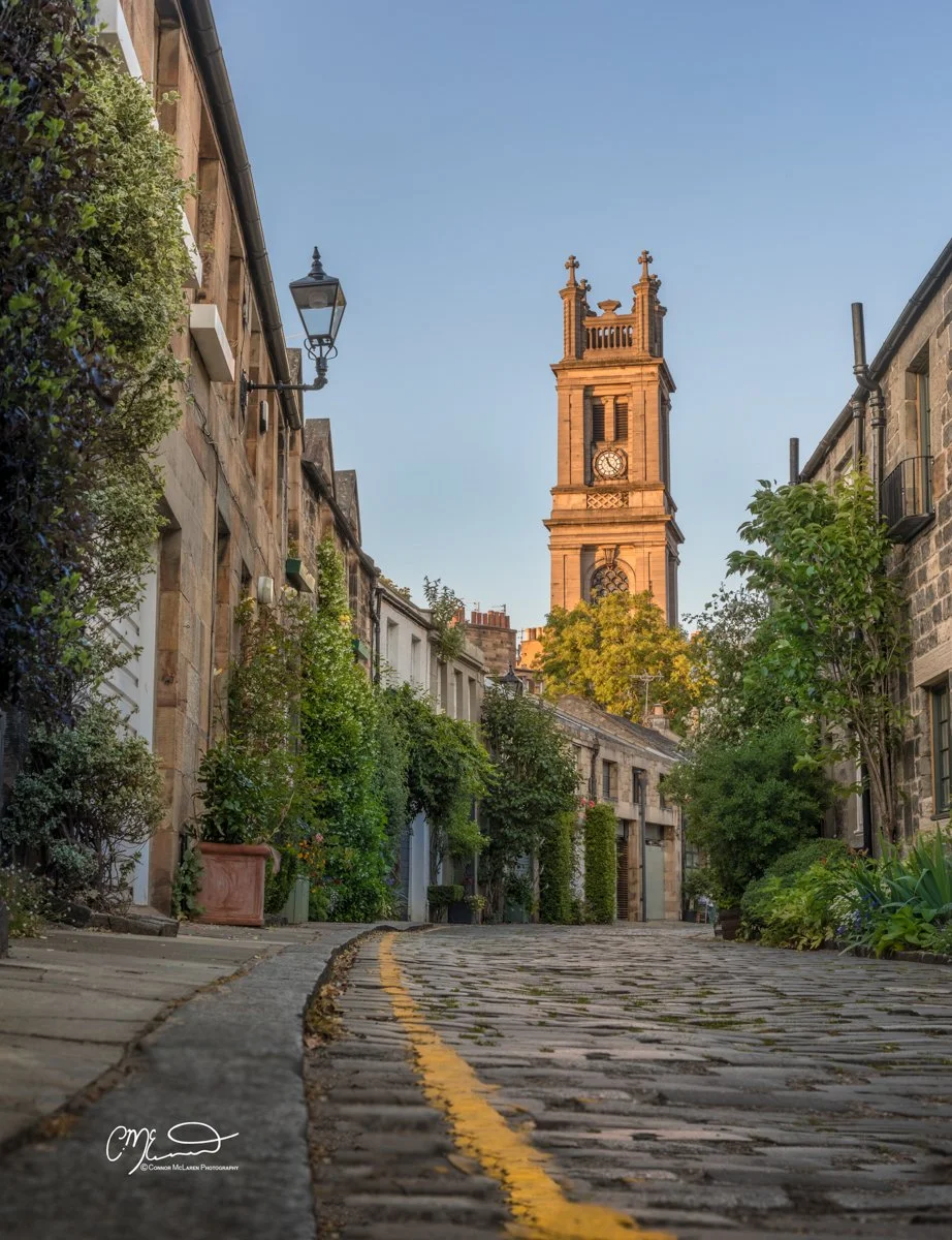 Scottish Lowlands, Scotland, Forth bridge Edinburgh. A cobblestone street lined with old stone buildings and greenery, leading to a tall clock tower with a pointed top, set against a clear blue sky.