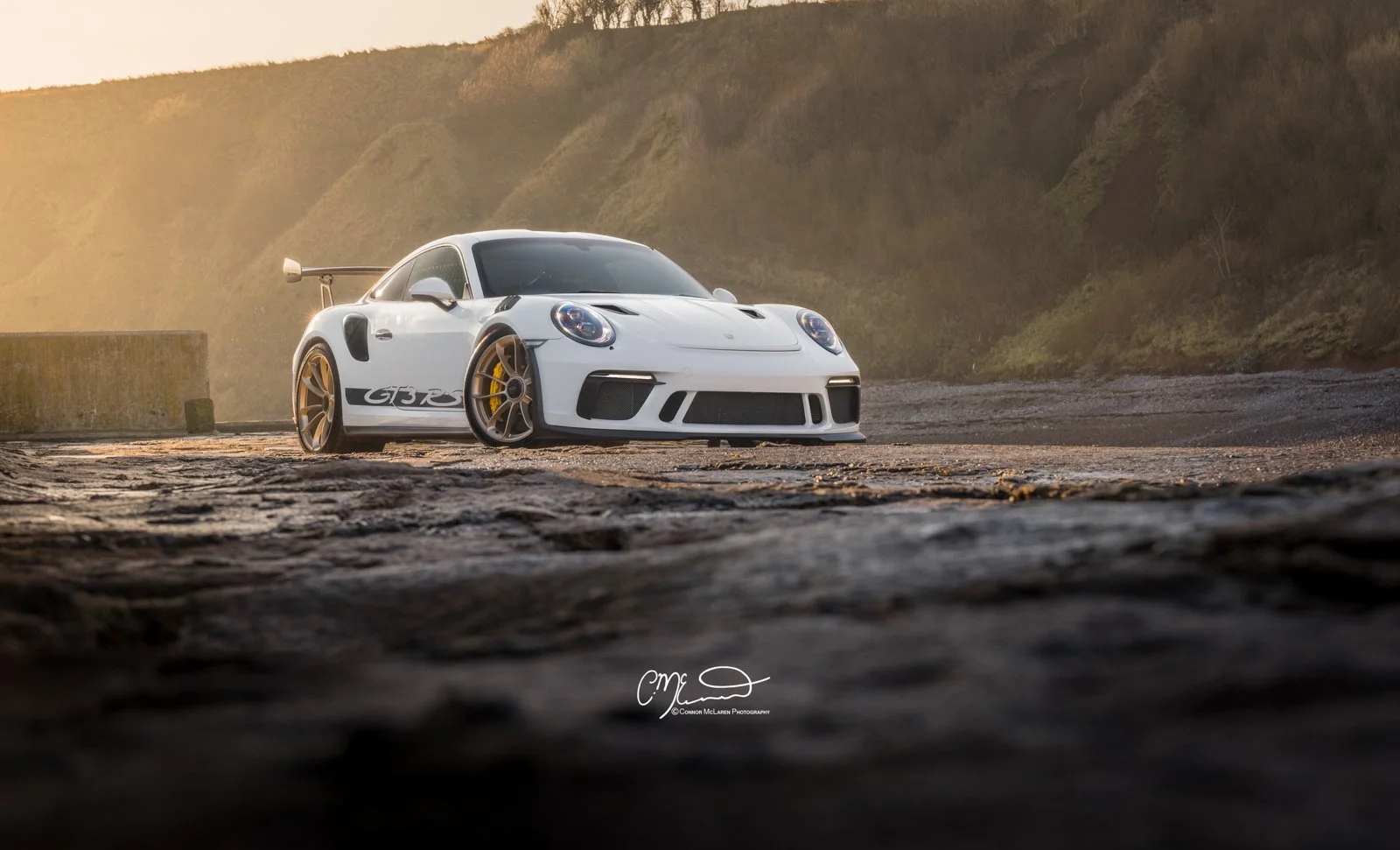 A white Porsche 911 GT3 RS race car with a large rear wing, detailed front bumper, and black side graphics, parked on a rocky coastal area with cliffs in the background during sunset.