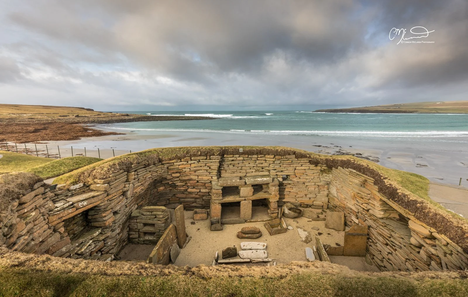 Skara Brae, Orkney, Scotland. Ancient stone ruins on a grassy hill overlooking a beach and ocean with a cloudy sky.