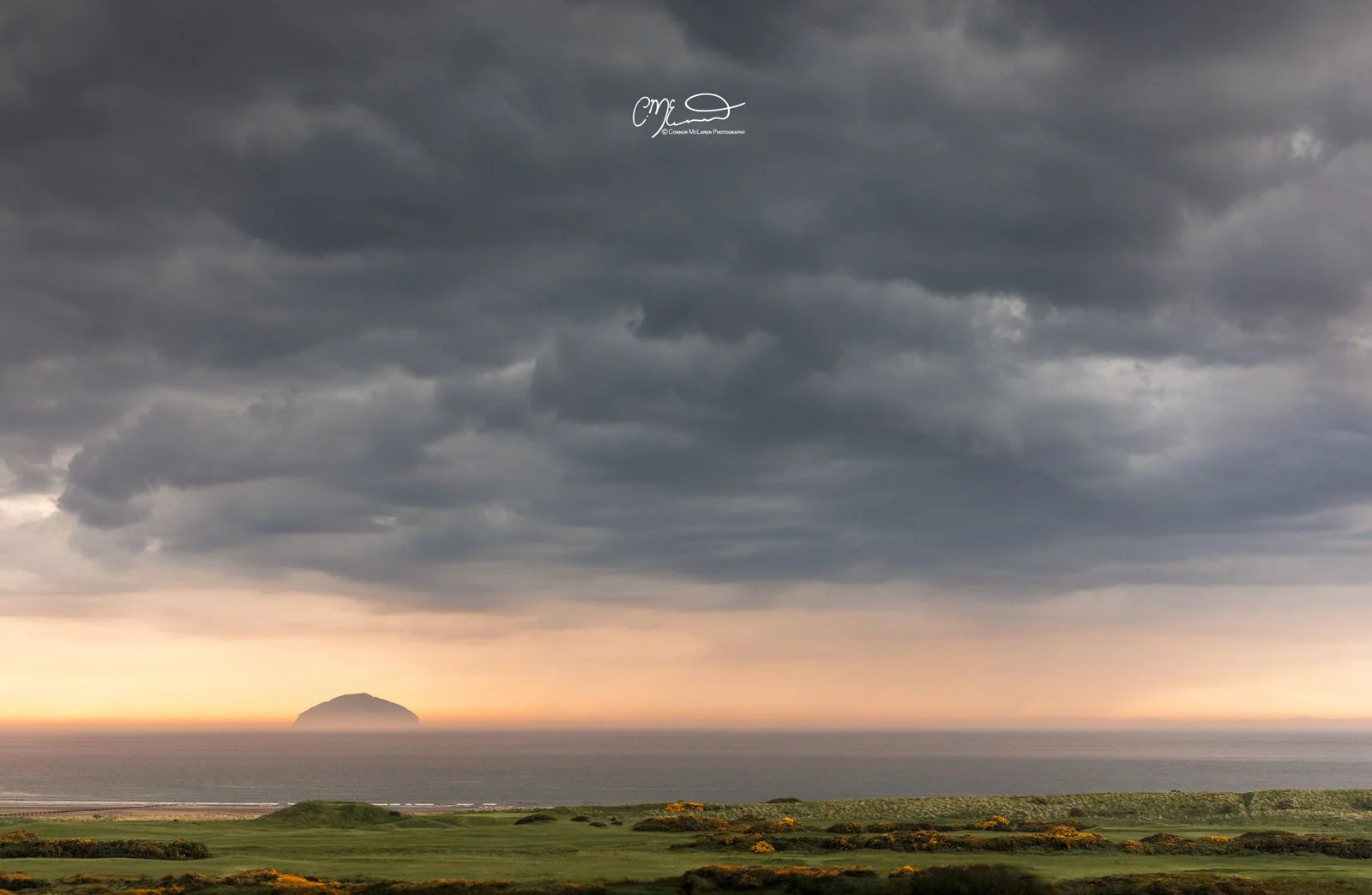Scottish Lowlands, Scotland, Turnberry. Dark storm clouds over the ocean with a small island on the horizon, and a green landscape with scattered bushes in the foreground.