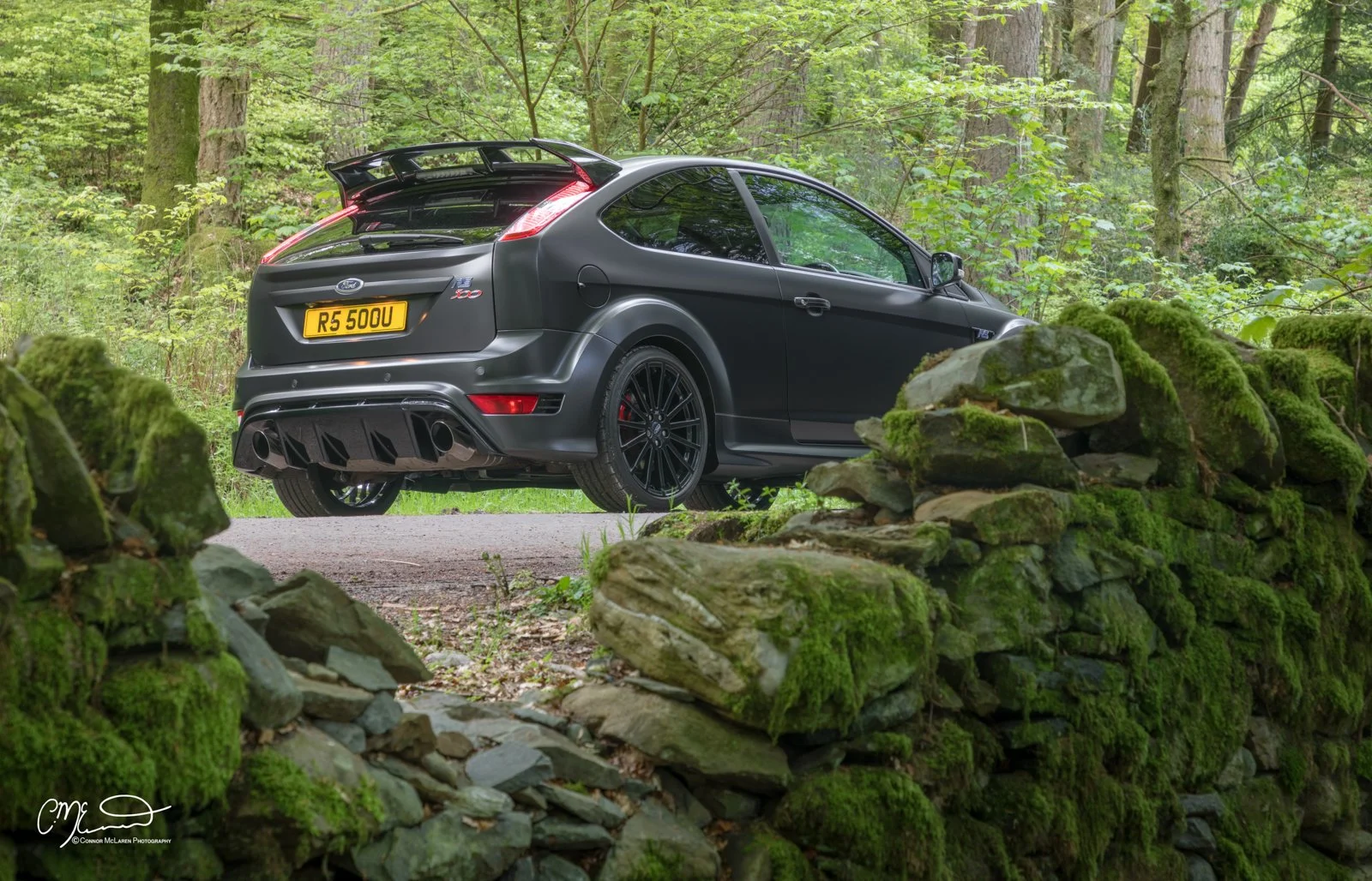 Ford Focus RS500. Black sports car with a spoiler and custom black rims parked on a forest road, surrounded by green trees and mossy rocks.