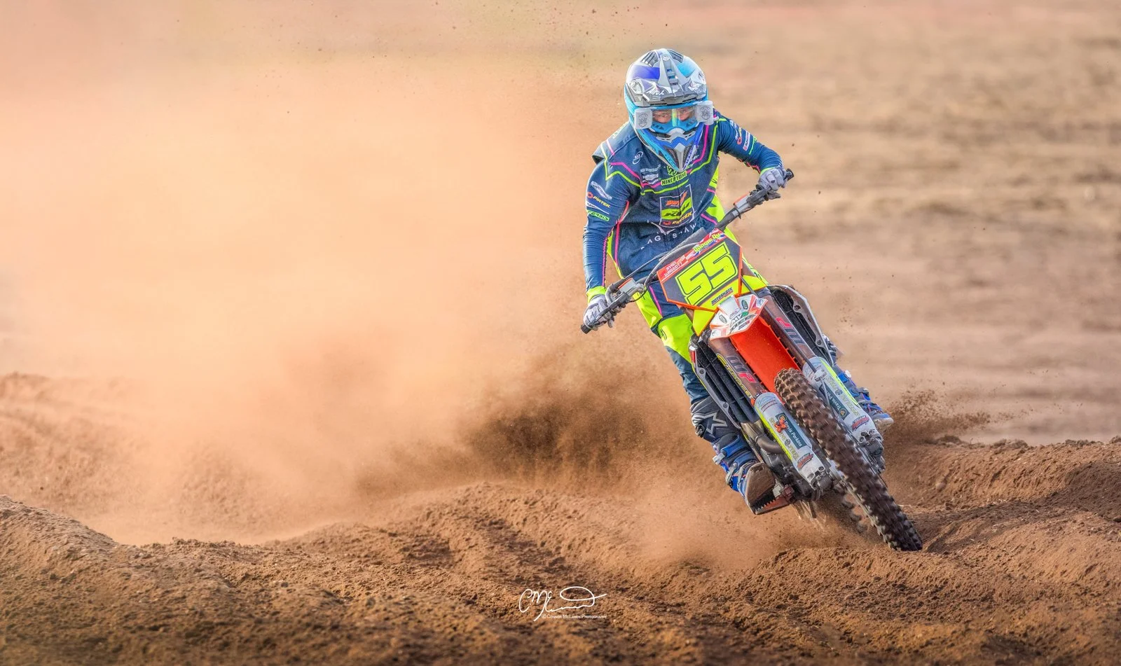 Motocross rider leaning into a turn on a dirt track, kicking up a cloud of dust, wearing a blue helmet and neon yellow and blue racing gear, number 55 on the bike.