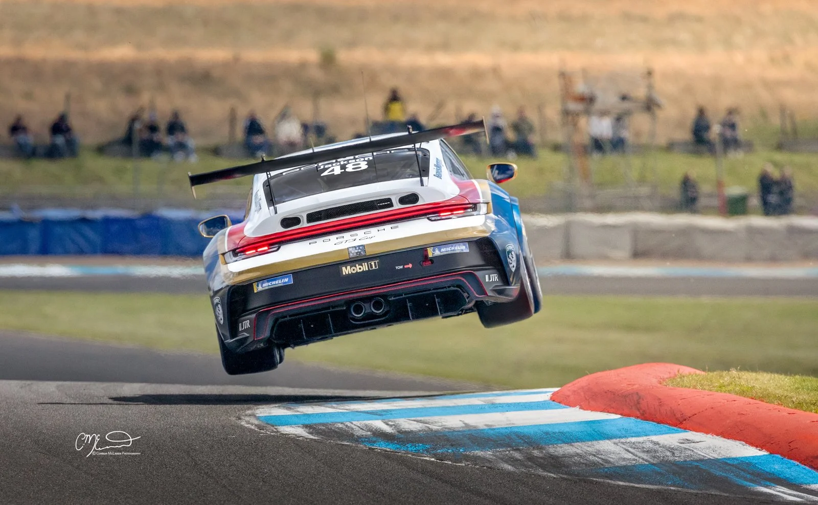 Porsche 911 GT3 Cup car racing in the Carrera Cup at Knockhill, takes off jumping through the air. A racing car, a Porsche 911, in mid-air after hitting a curb on a race track, with spectators in the background.