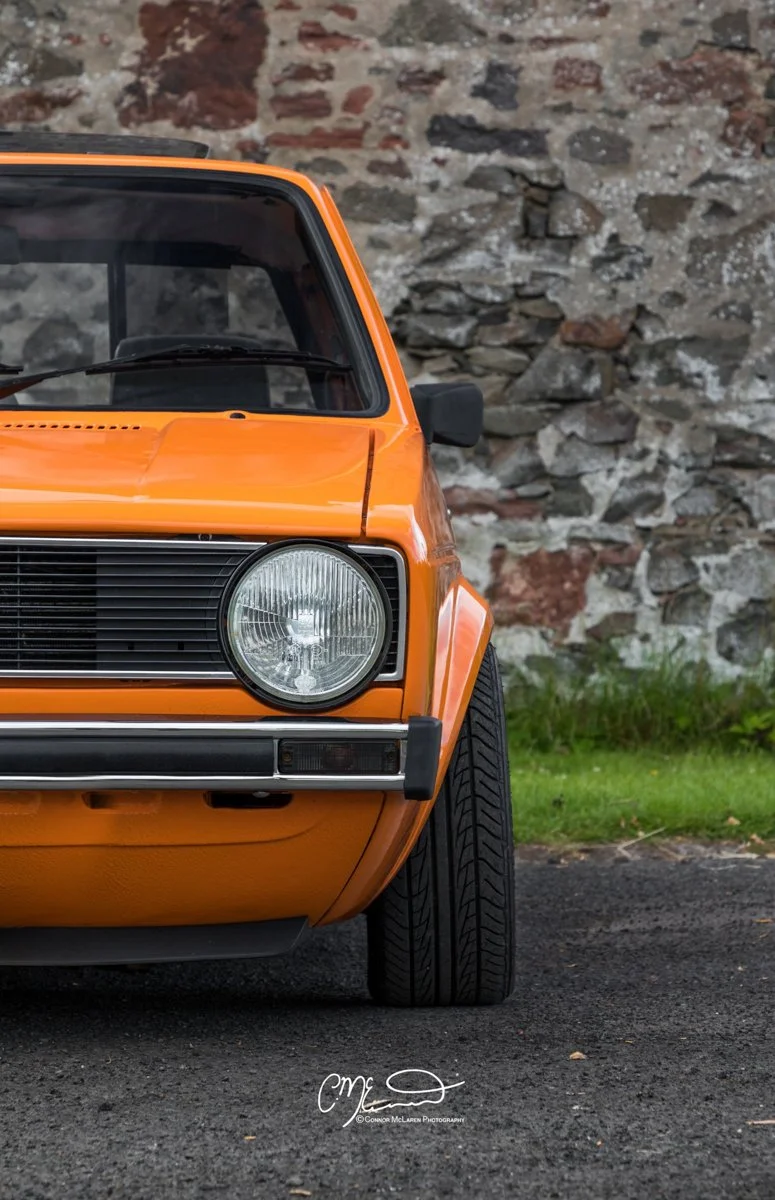 Orange vintage Volkswagen Caddy pickup truck Close-up of the front left side of an orange vintage car parked on a dark asphalt surface, with a stone wall in the background.