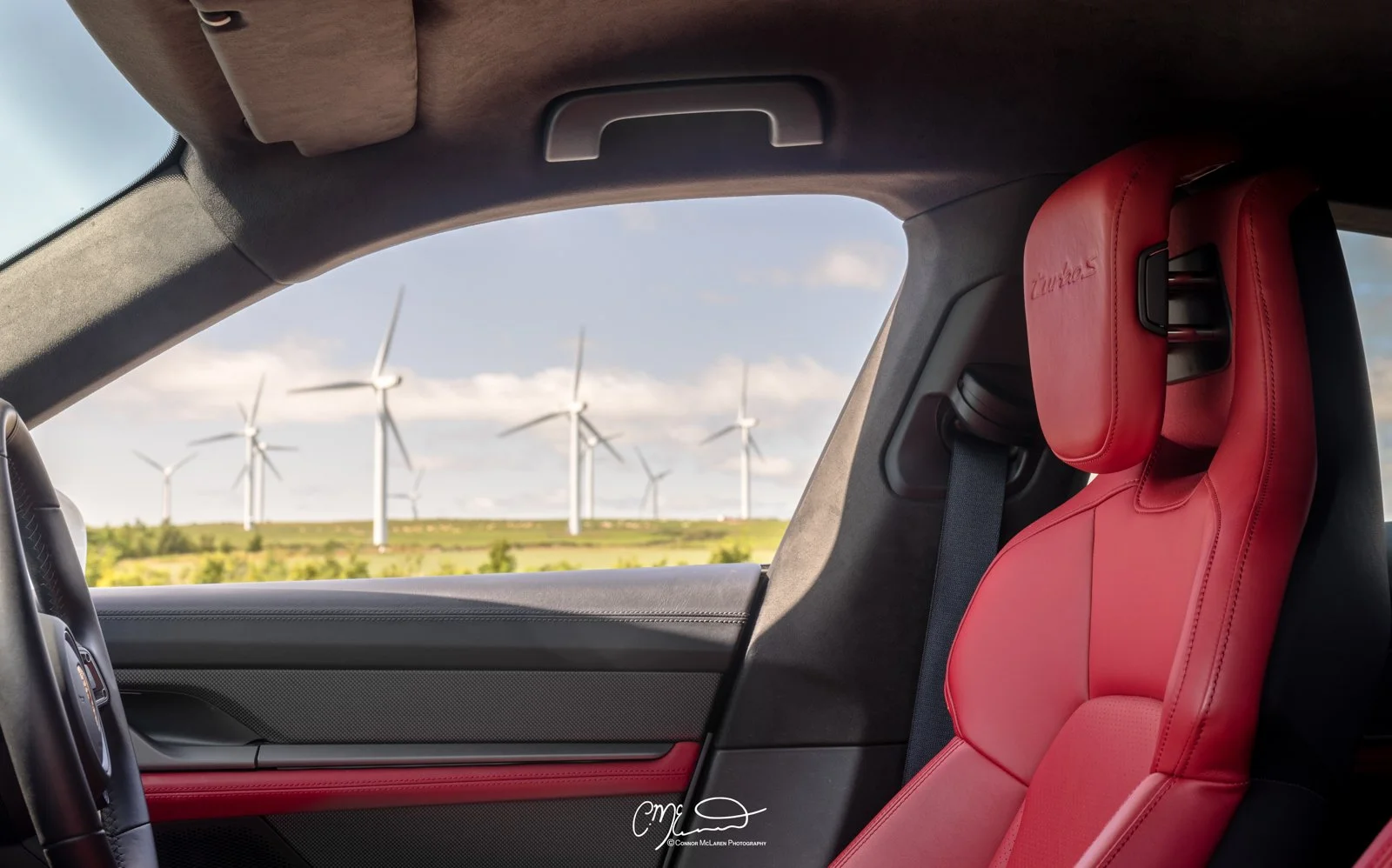 Porsche Taycan red Interior, Turbo S. Interior of a car with a red seat, window view of wind turbines in a green landscape under a partly cloudy sky.