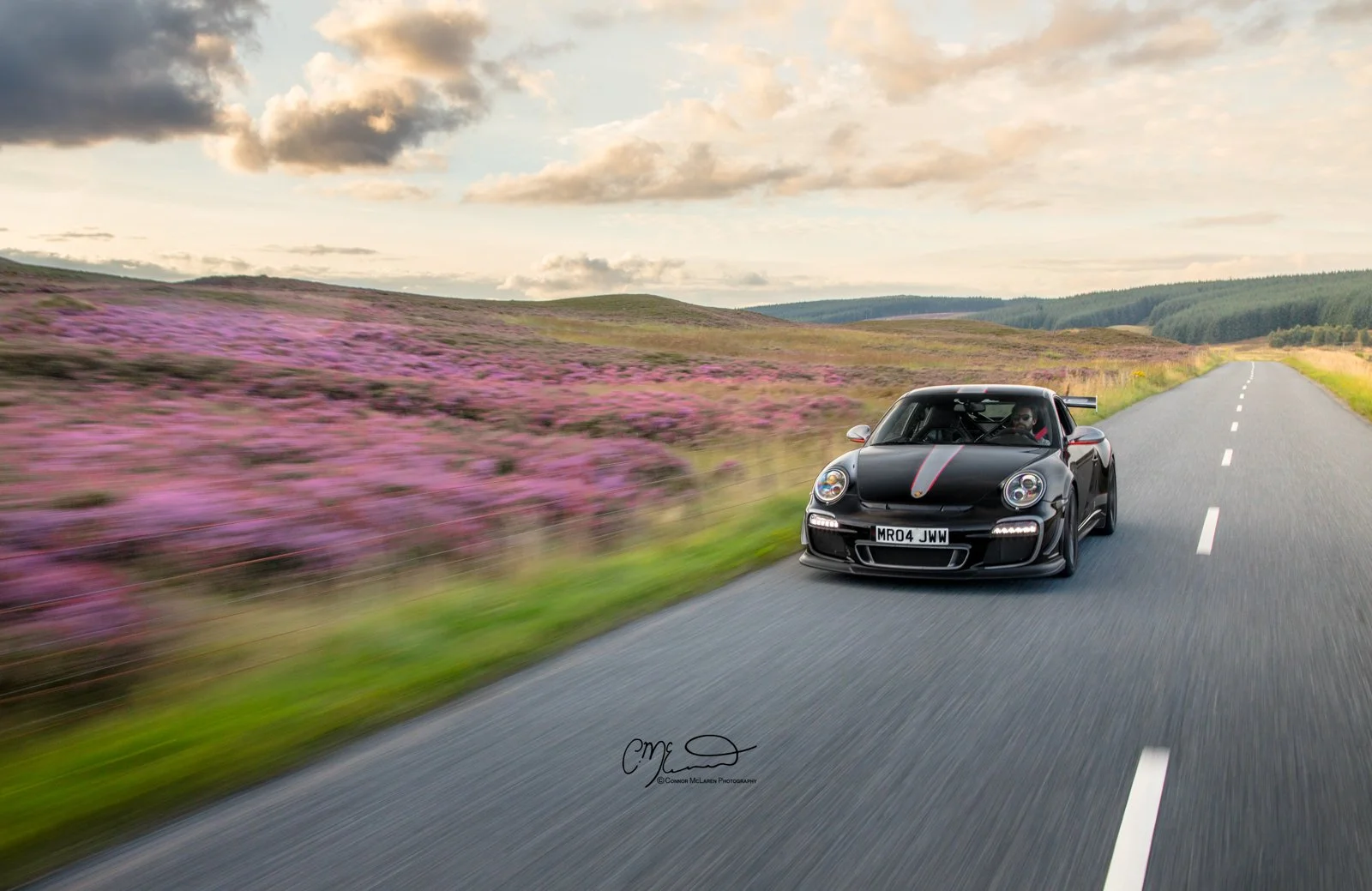 Porsche 911 GT3 RS 4.0L. A black sports car with a red stripe driving on an open road through a scenic landscape with purple heather and rolling hills under a partly cloudy sky.