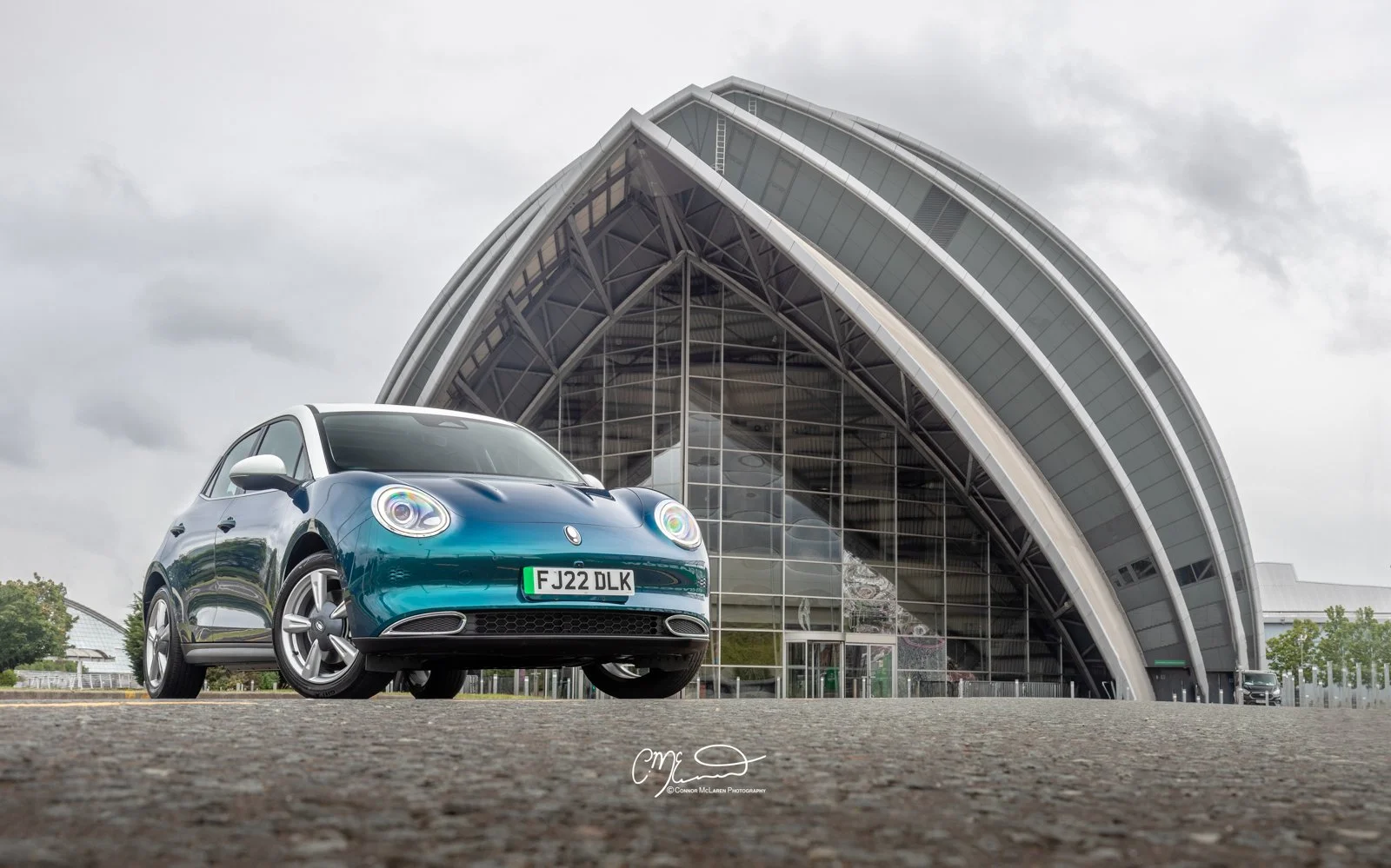 A blue Ora Funky Cat electric car parked in front of a modern, arched glass building on a cloudy day.